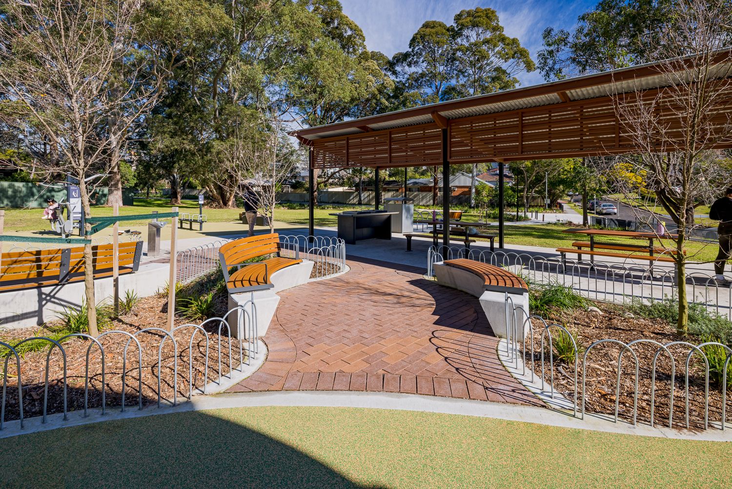 Shaded park seating area with a pergola roof, curved timber benches and brick paving, beside a playground and green lawn.