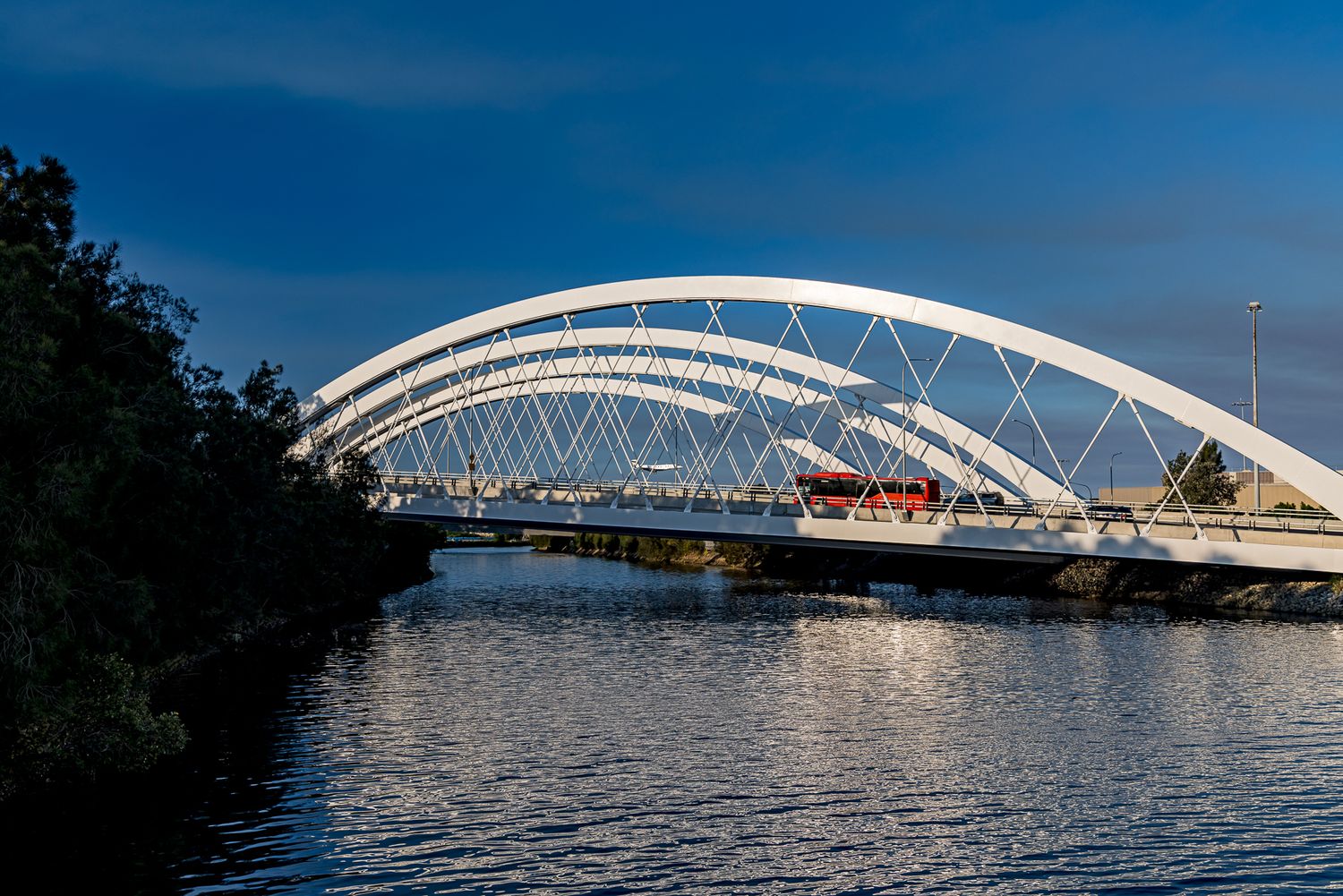 White arched bridge with a red vehicle crossing over a still river reflecting the soft blue sky.