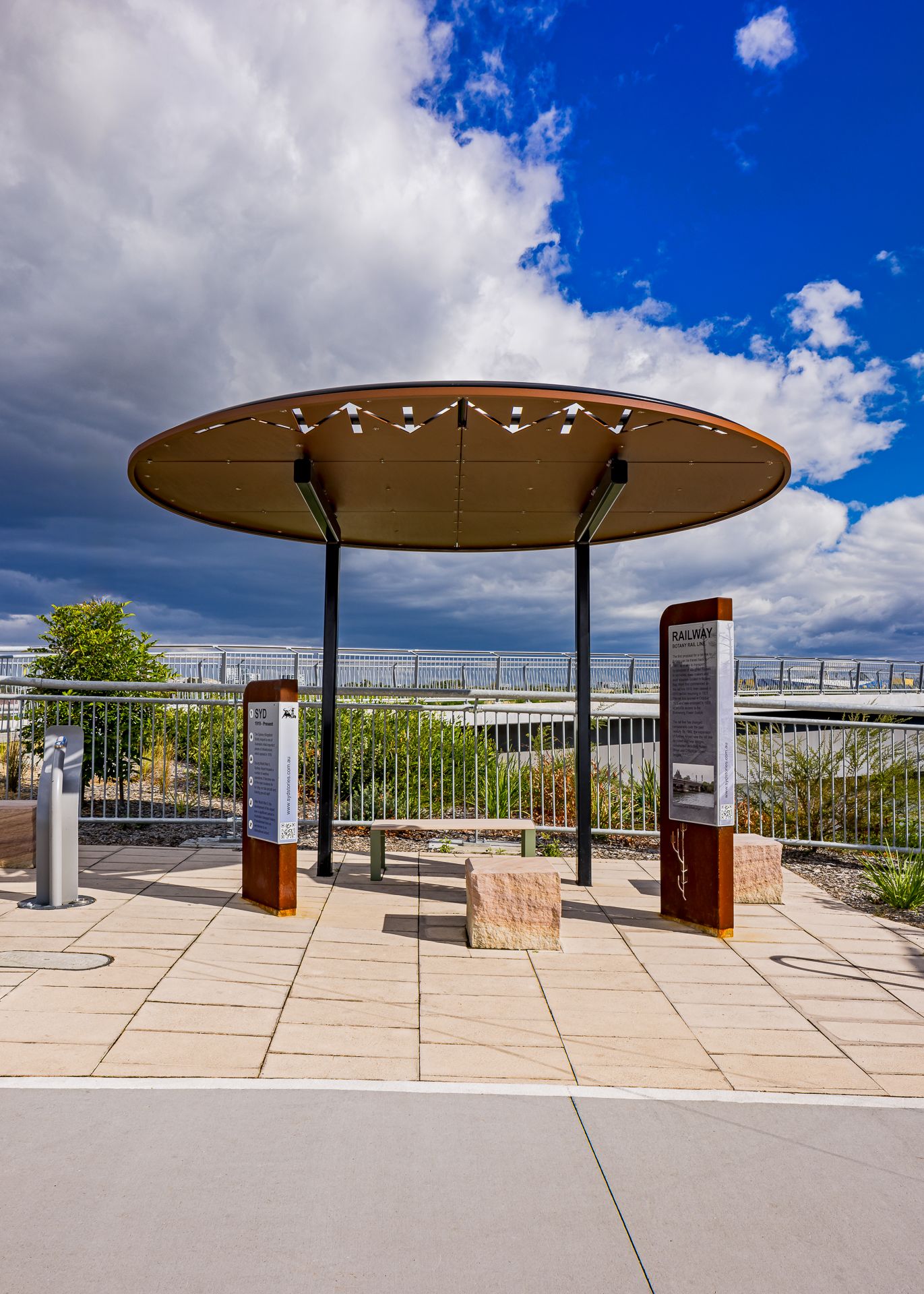 Circular roofed rest area with stone seating and interpretive signs overlooking green plants and a railing under bright blue sky and clouds.