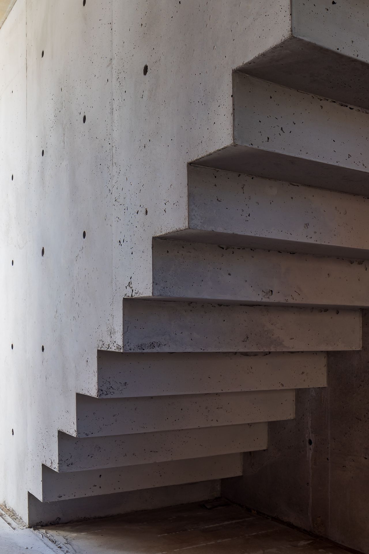 Cantilevered concrete staircase with exposed steps and visible tie holes along a raw concrete wall.