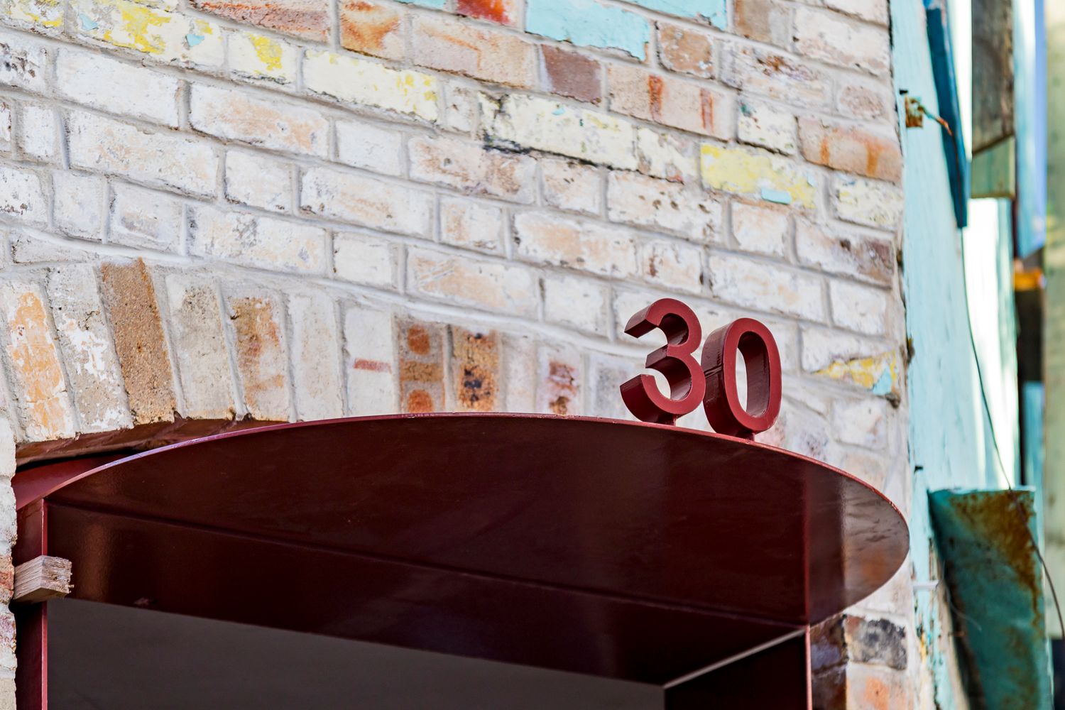Red metal house number 30 mounted on a weathered painted brick wall above a curved dark red canopy.
