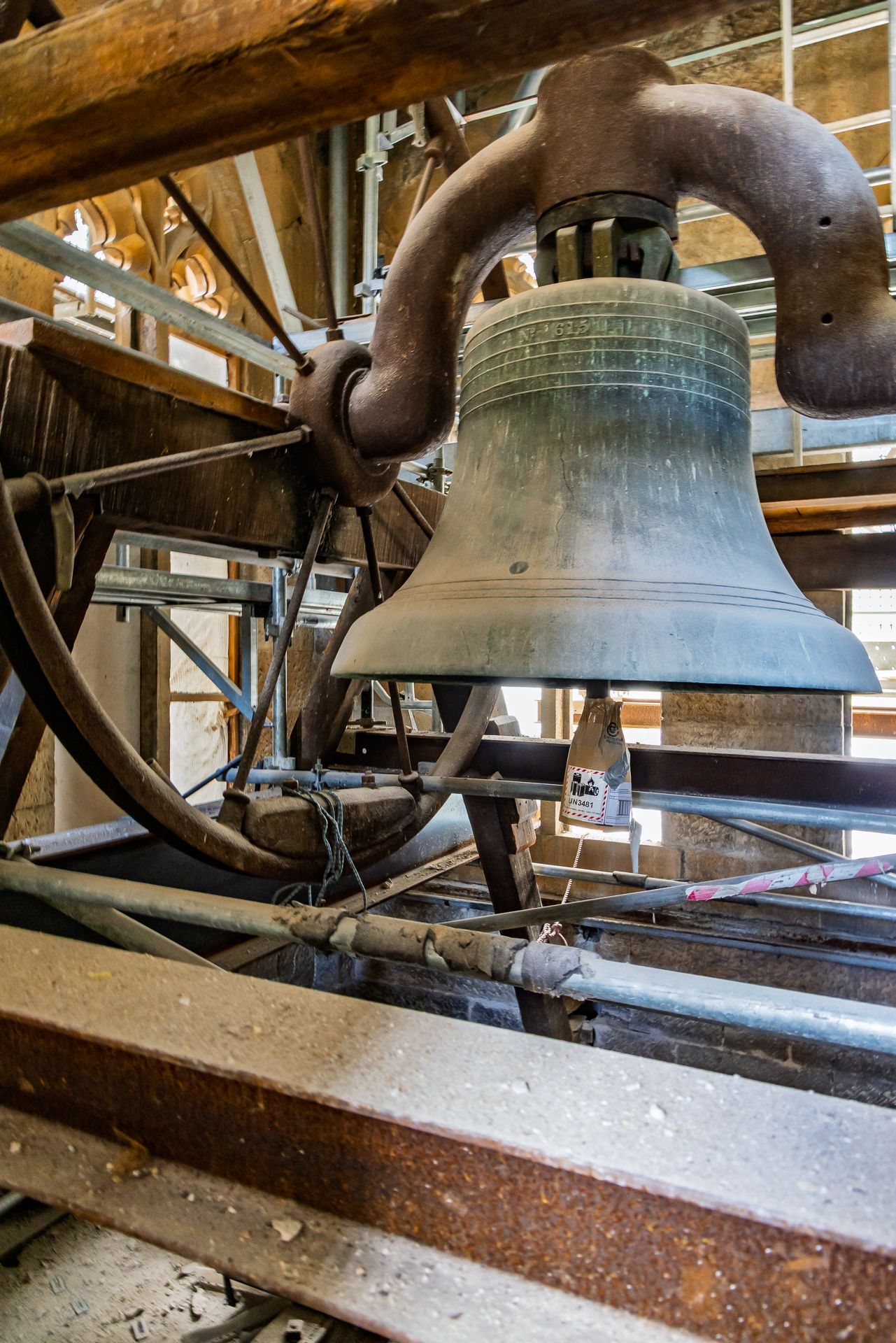 Large bronze church bell hanging from a wooden frame inside a bell tower with metal pipes and beams.