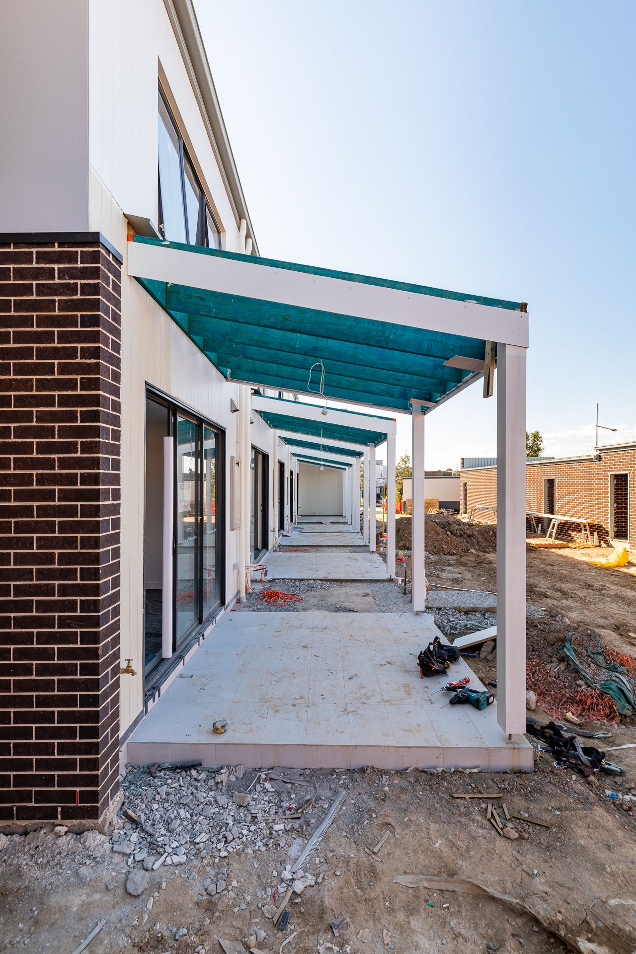 New townhouse walkway with brick feature wall, sliding glass doors and teal covered porch roofs on a construction site.