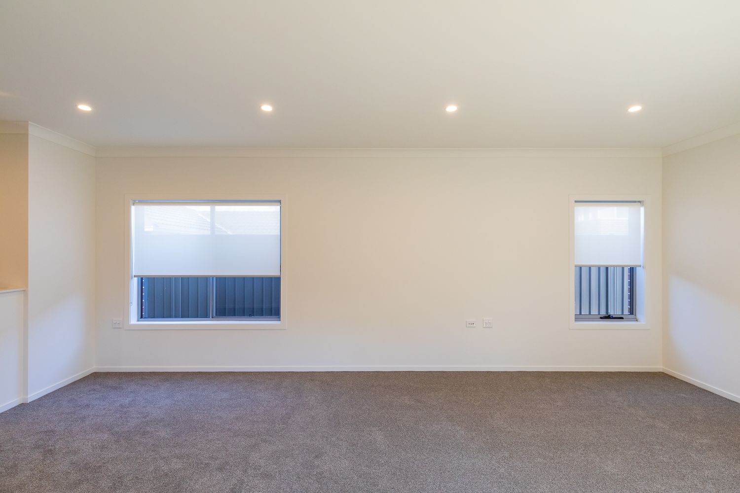 Empty open-plan living room in a new home with fresh white walls, grey carpet, recessed ceiling lights and two windows with blinds.