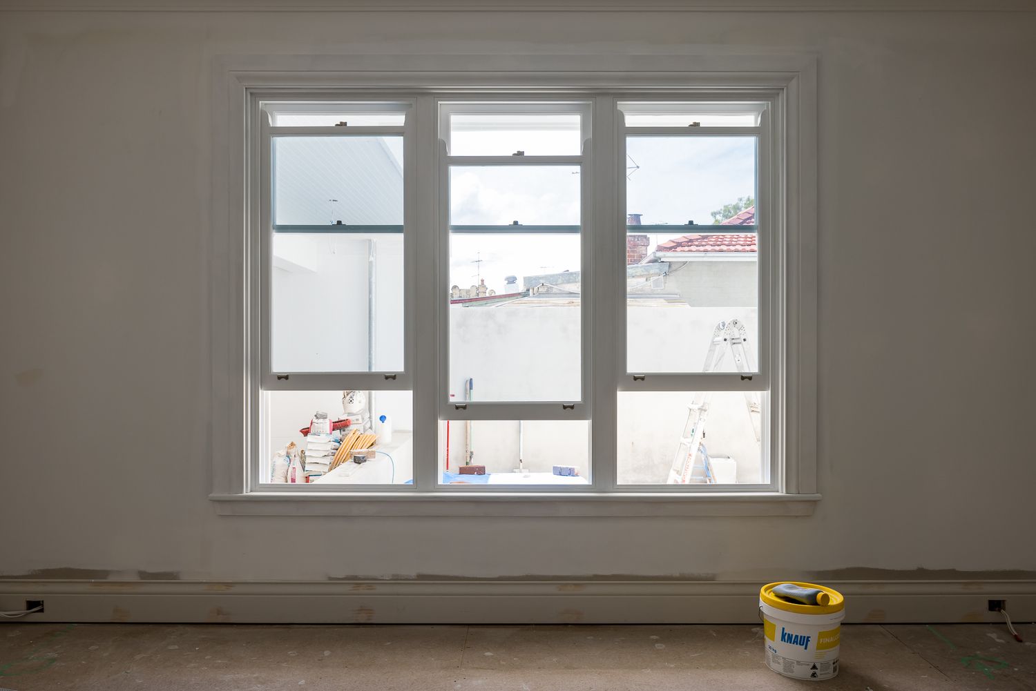 Triple white-framed window in a room under renovation, with a paint bucket on the floor and rooftops and sky visible outside.