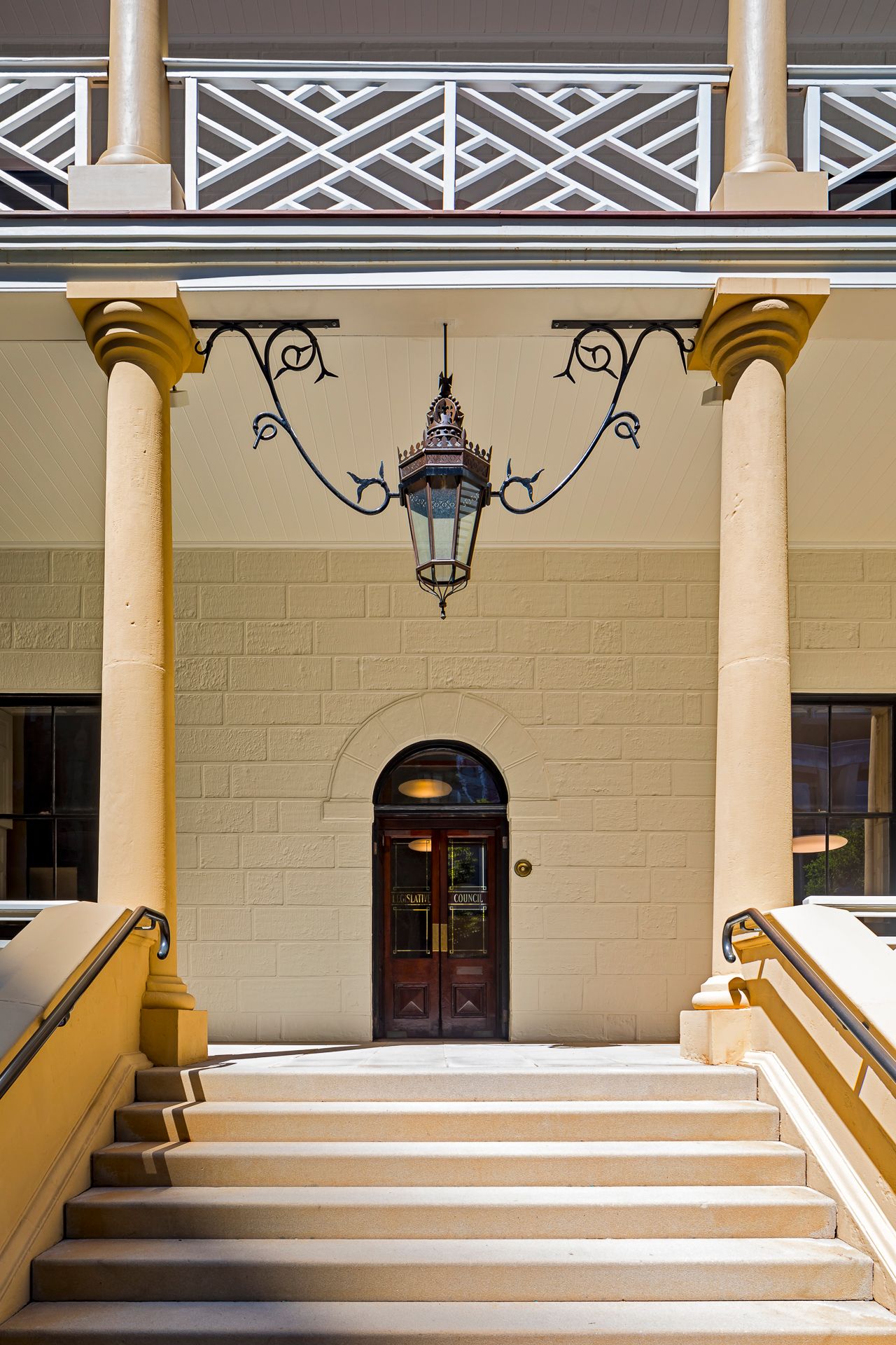 Sandstone building entrance with a wrought iron hanging lantern, tall columns, arched wooden doors and wide steps leading up to a balcony.