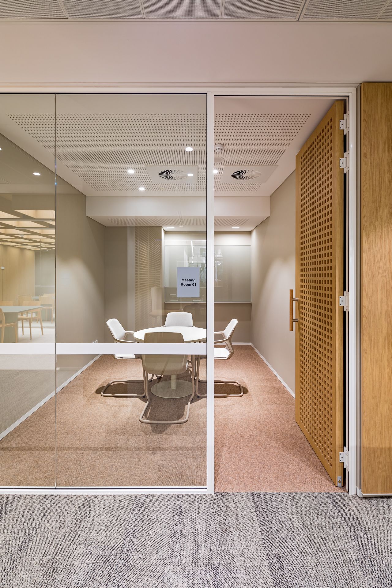 Small glass-walled office meeting room with a round white table, modern chairs and an open perforated wooden door.