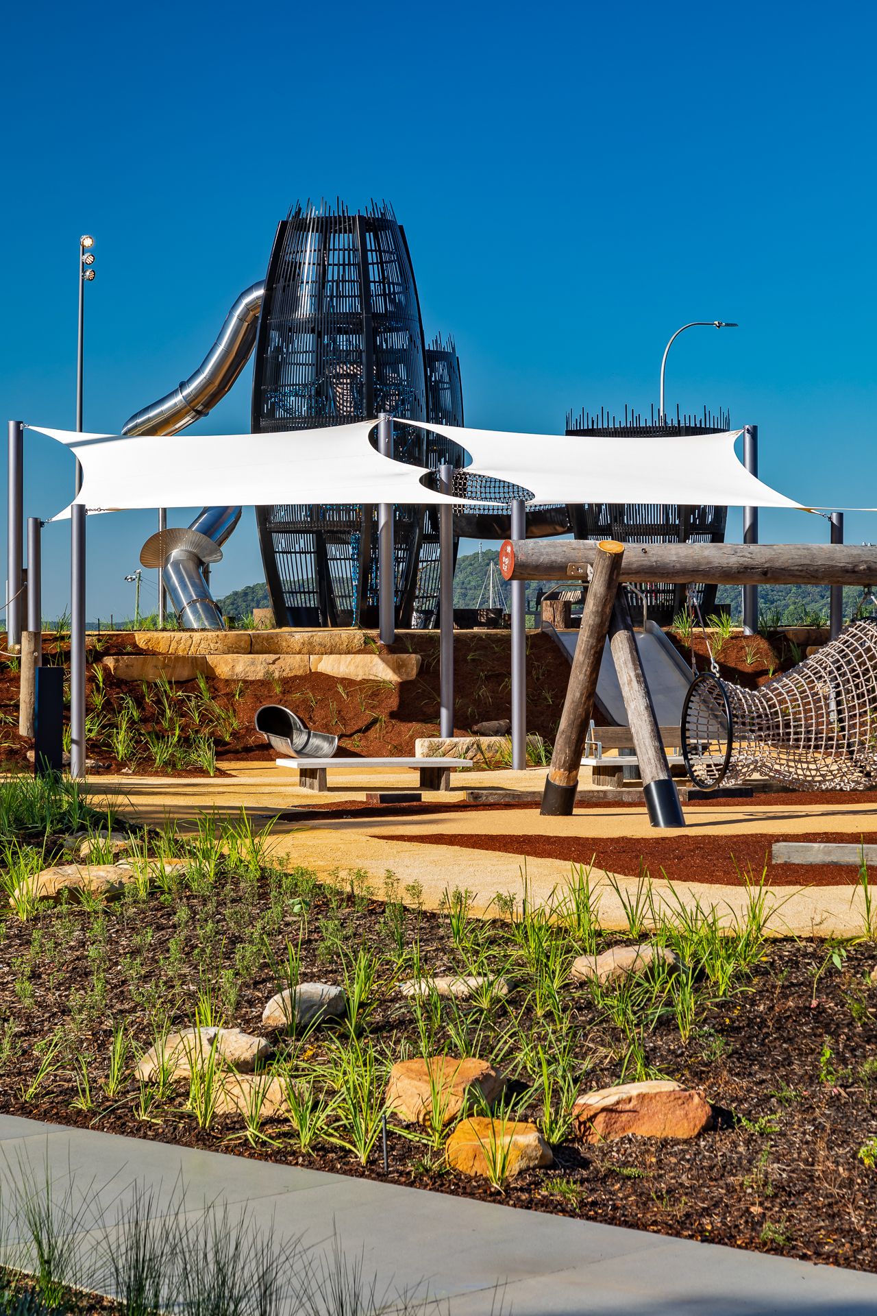 A playground with shade sails, rope net swings and a lattice slide tower sits behind native plantings and sandstone rocks.