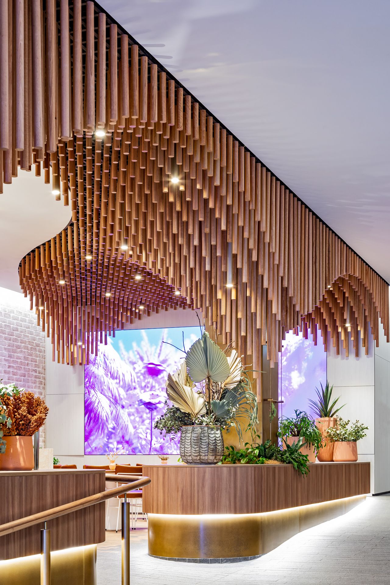 Sweeping timber tube ceiling above a curved wooden counter styled with potted plants, with a vibrant purple tropical mural behind.