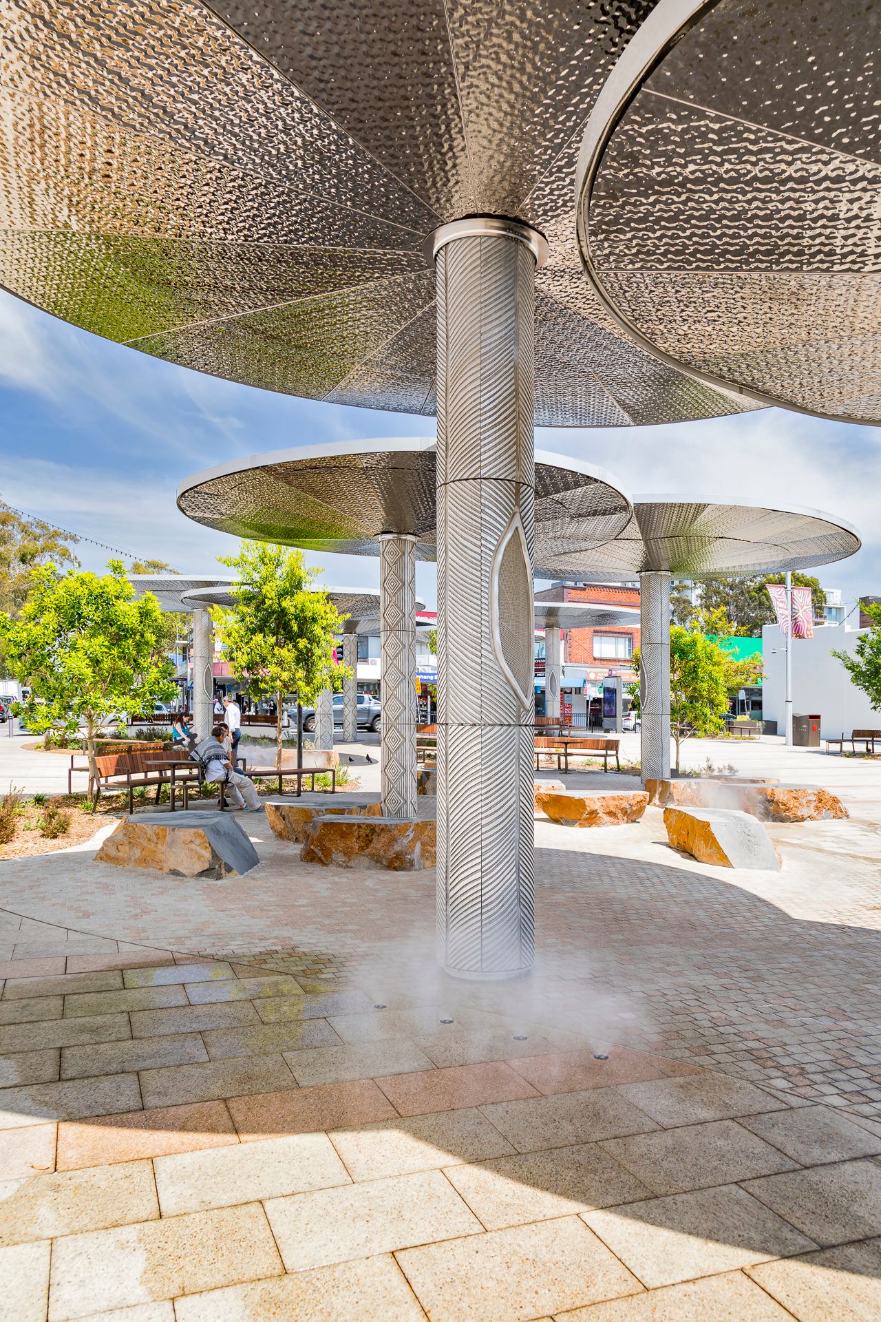 Mist rises around a patterned steel column beneath large canopies in a sunlit plaza with sandstone seating and brick paving.