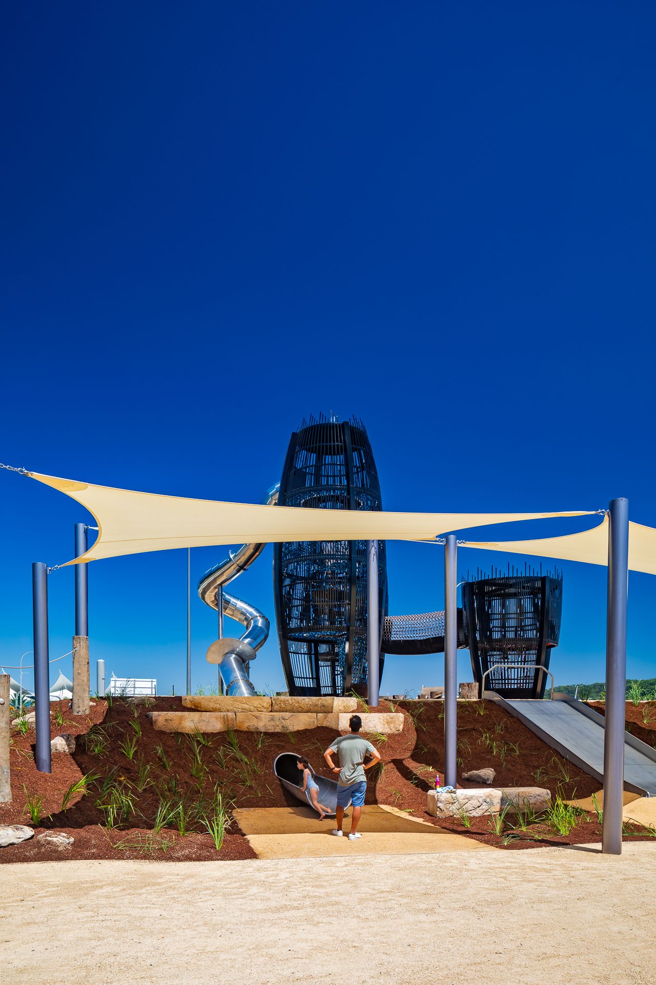 Visitor and child at a hillside playground with a tall metal slide tower and cream shade sails under a bright blue sky.