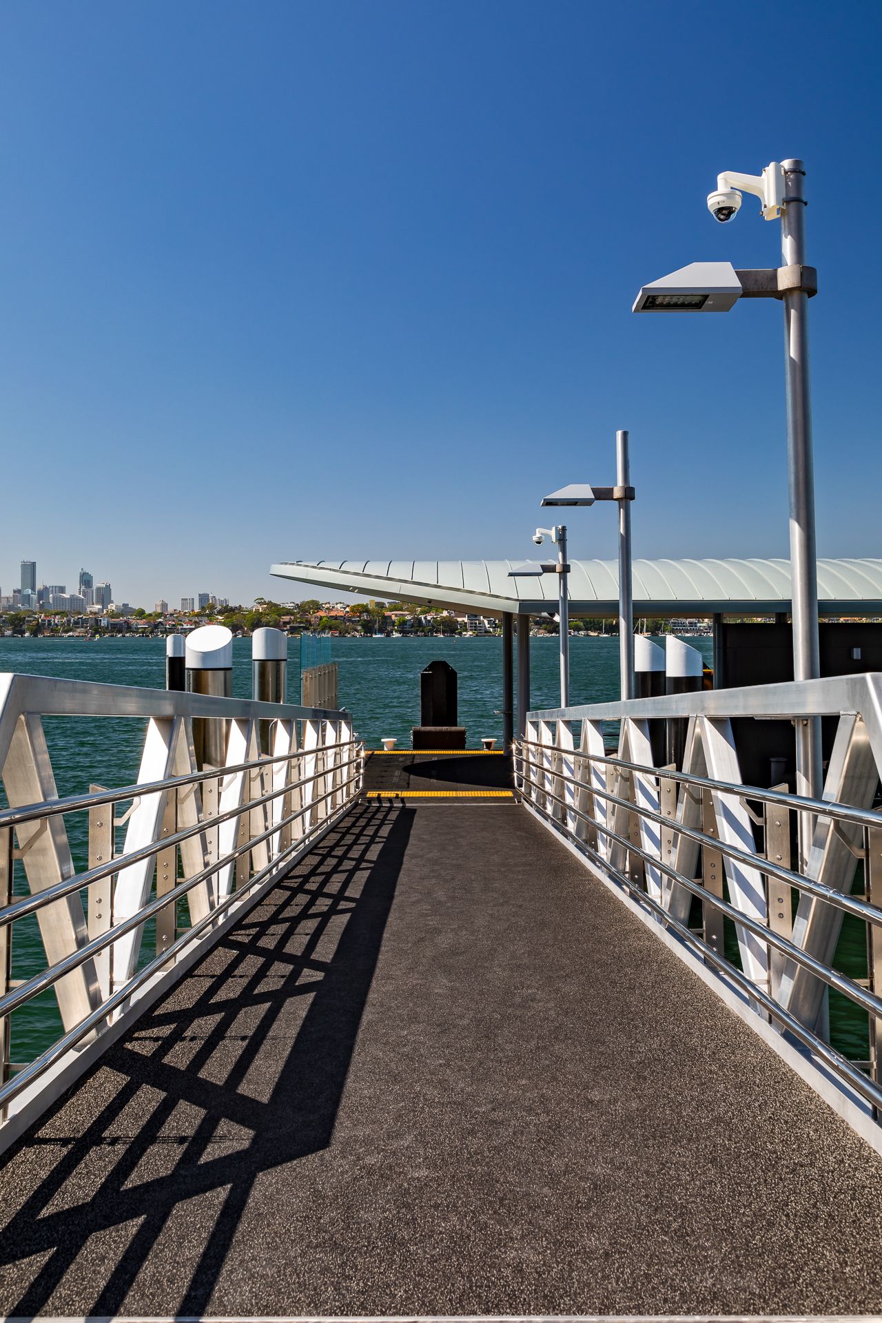 Empty ferry wharf walkway with metal railings and security cameras leading to a covered pontoon, with a city skyline beyond.