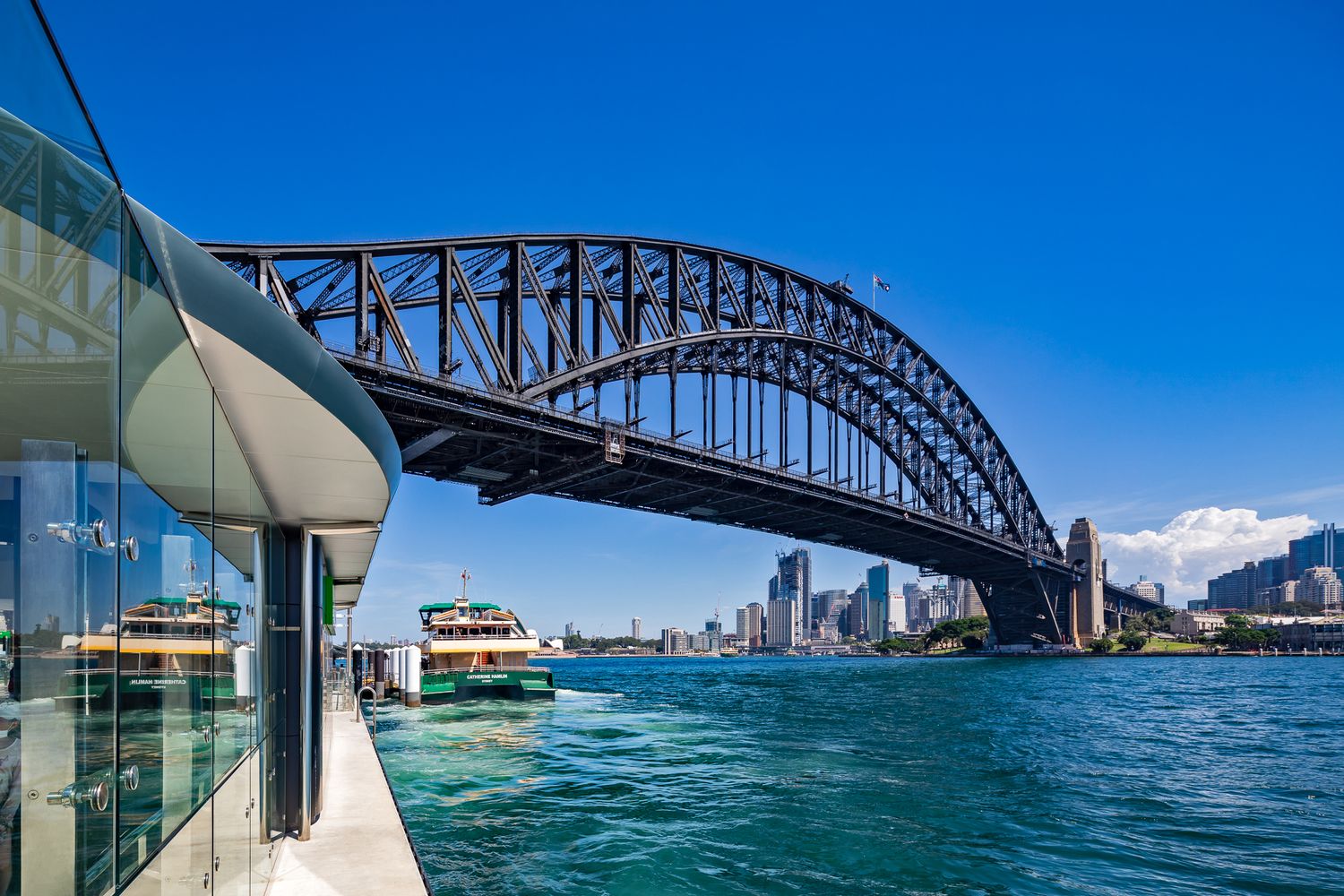Sydney Harbour Bridge arches over blue water as a ferry docks beside a glass-fronted wharf with the city skyline beyond.