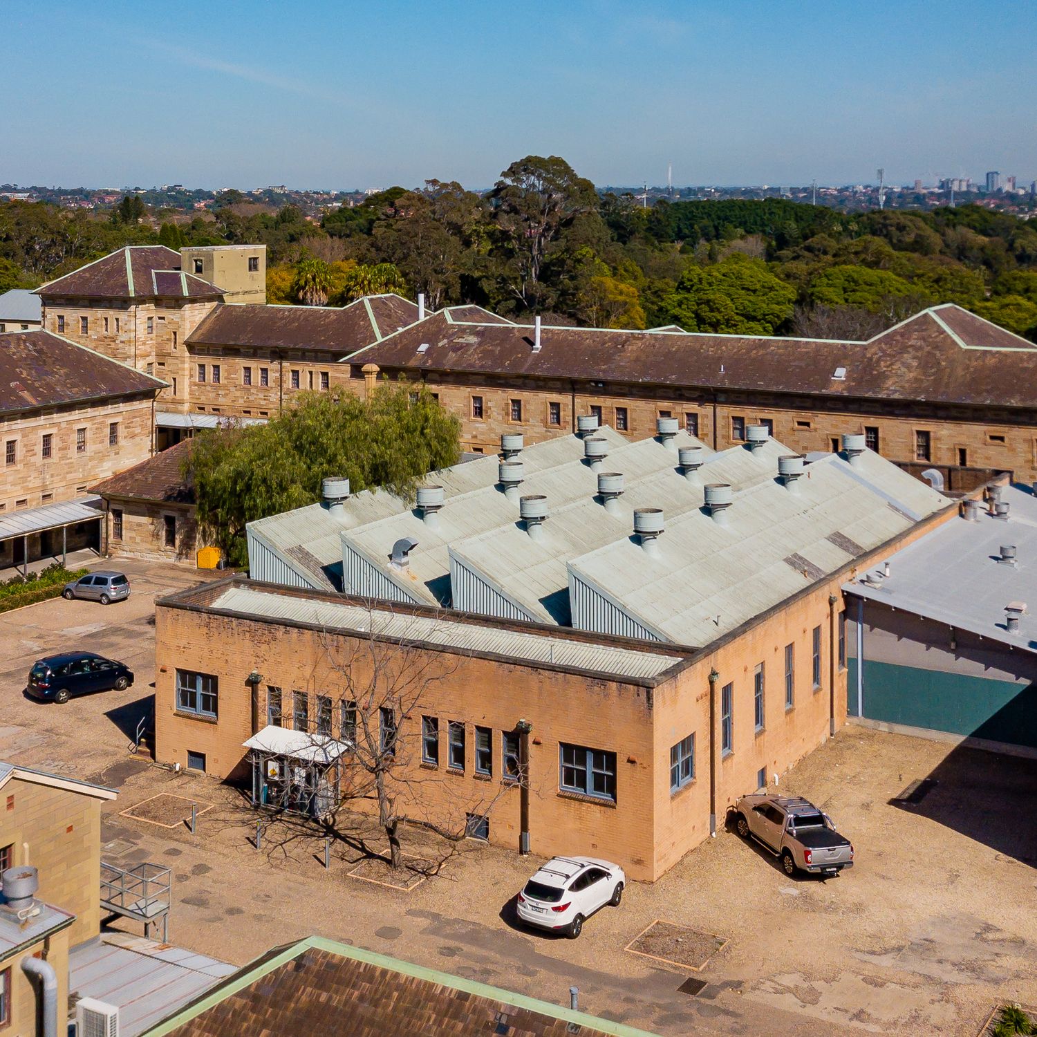 Aerial view of historic brick buildings with a sawtooth roof workshop, parked cars and leafy parkland beyond.