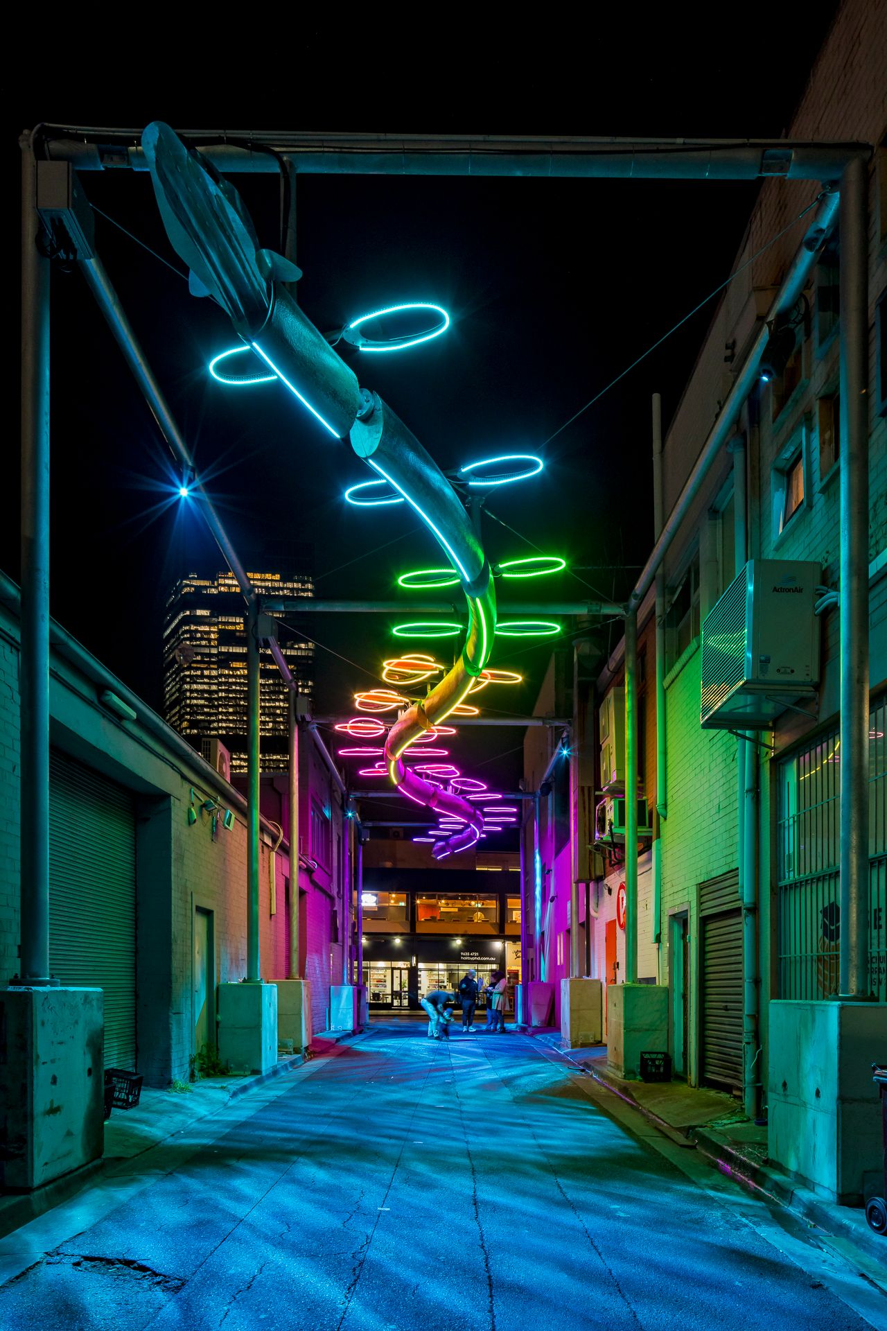 Colorful neon fish sculpture with glowing rainbow rings suspended above a dark urban alleyway at night, city skyscraper in the distance.