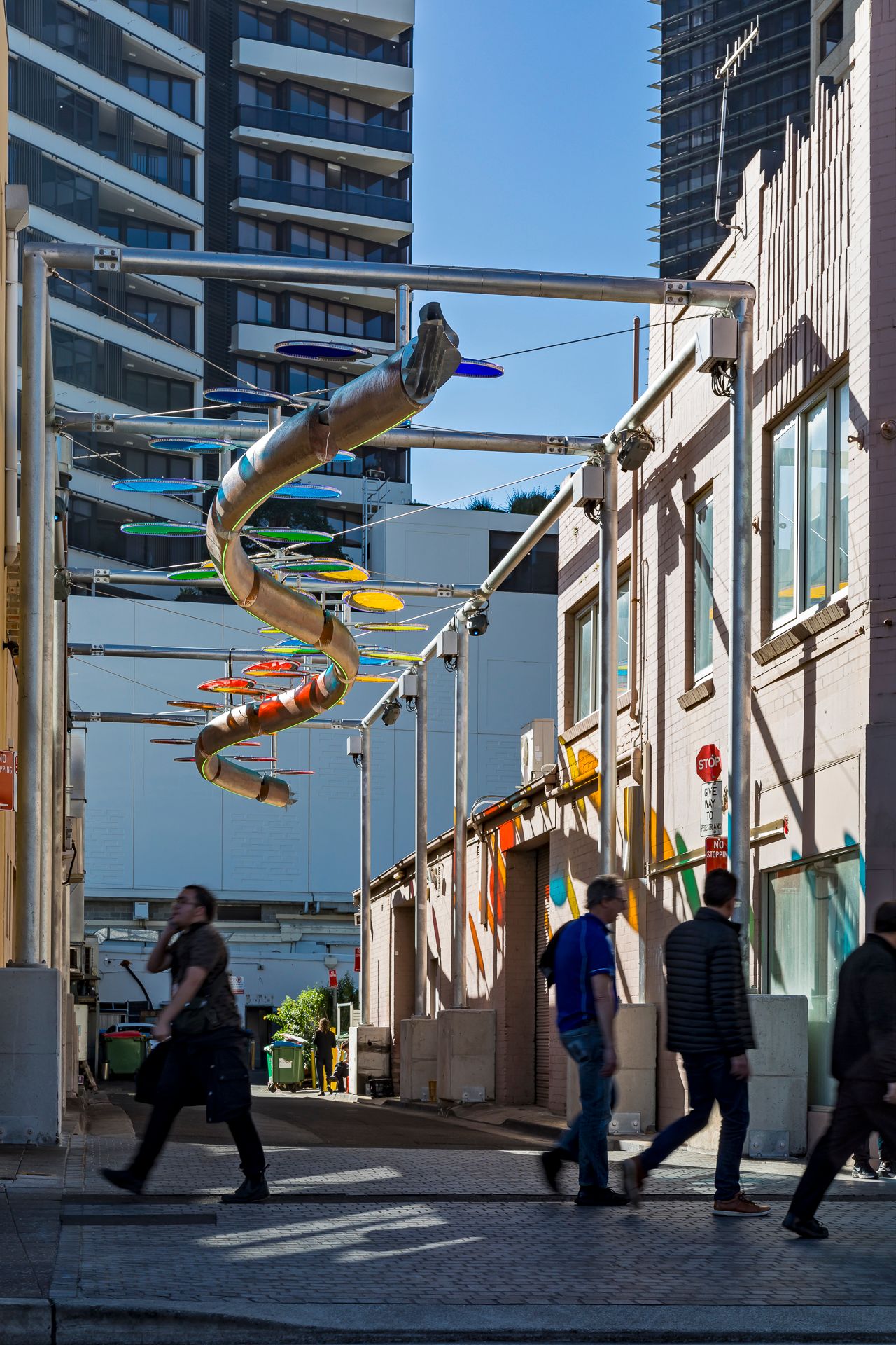 Pedestrians walk through a city laneway beneath a spiraling sculpture of colorful discs, with apartment towers behind.