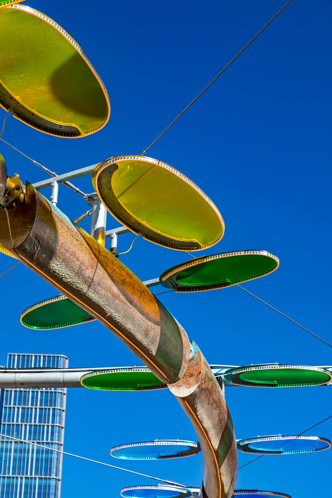 Large yellow-green and blue discs on a copper-toned curved sculpture against a saturated blue sky and glass tower.