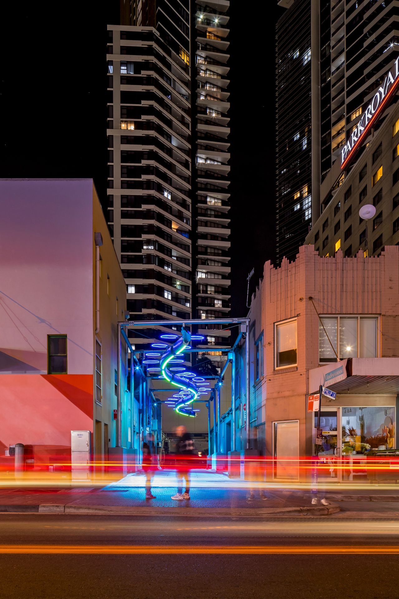 Blue and green neon spiral artwork lights a narrow city laneway at night with red light trails and tall apartment towers above.