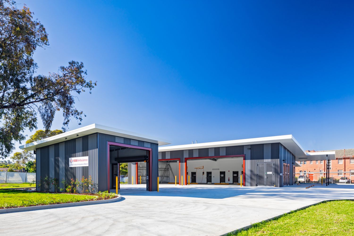 Modern single-storey building with dark gray cladding, red accents and a covered entrance beside landscaped lawn under a clear blue sky.