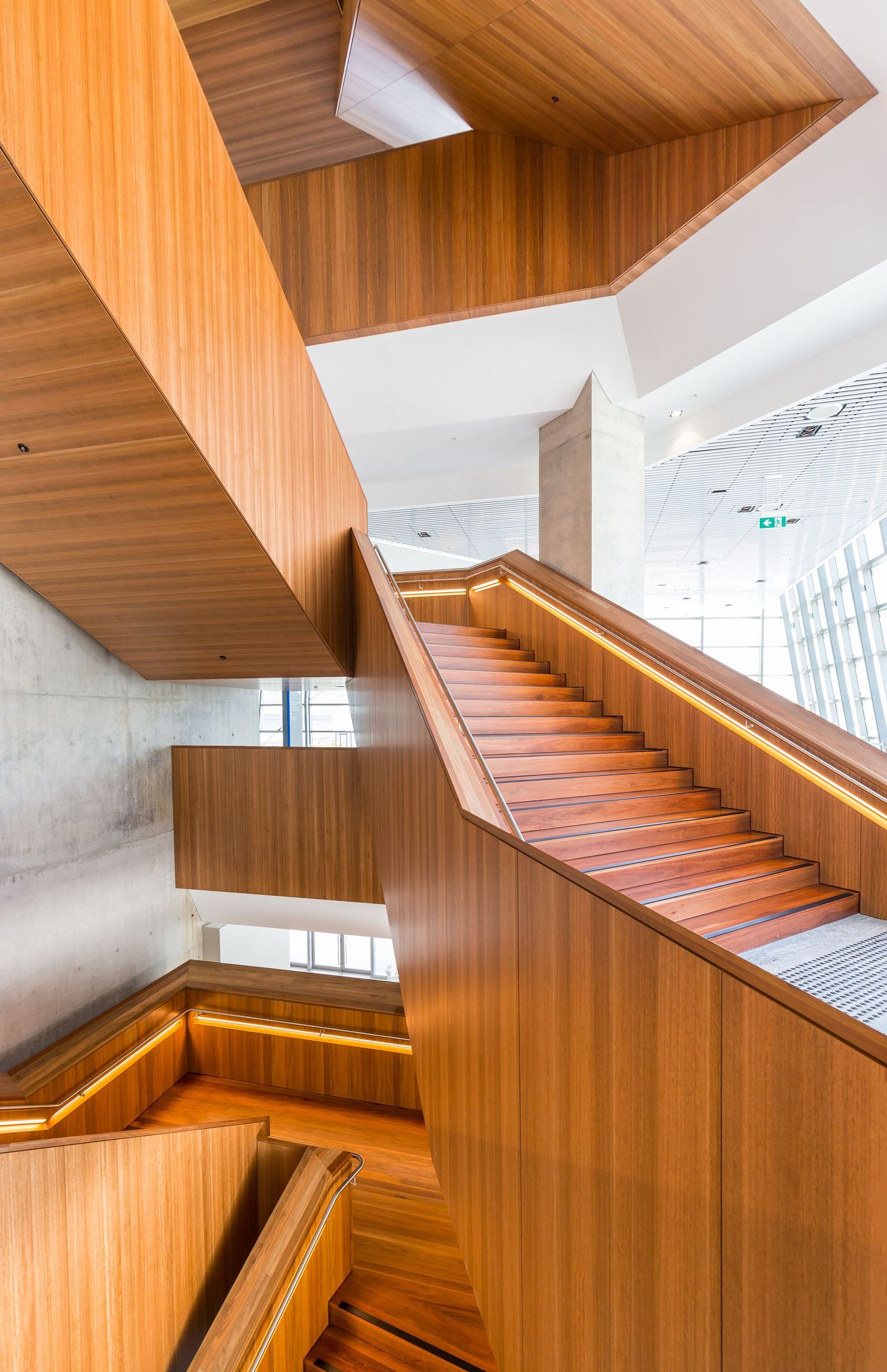 Multi-level timber-clad staircase with warm lighting, concrete walls and angular wood panelling inside a modern building.