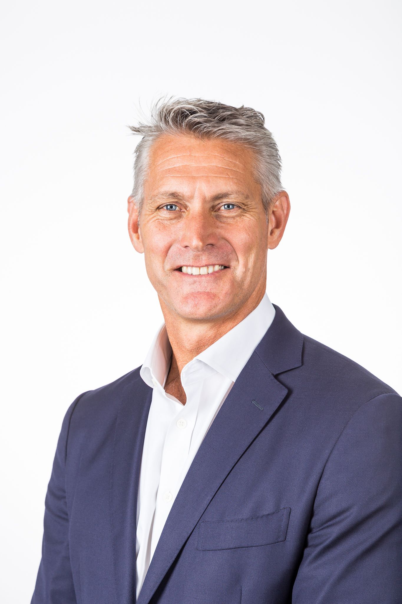 Smiling man with short gray hair wears a navy blazer and open-collared white shirt in a professional headshot against a white background.