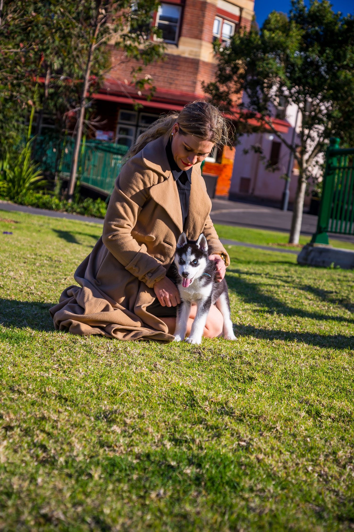 A woman in a long camel coat kneels on a grassy lawn beside a husky puppy, with brick buildings and trees behind.
