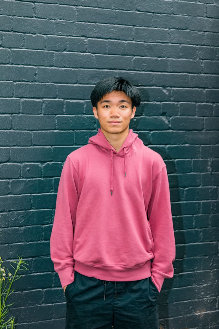 Young man with dark hair wearing a pink hoodie and navy pants stands against a dark painted brick wall.