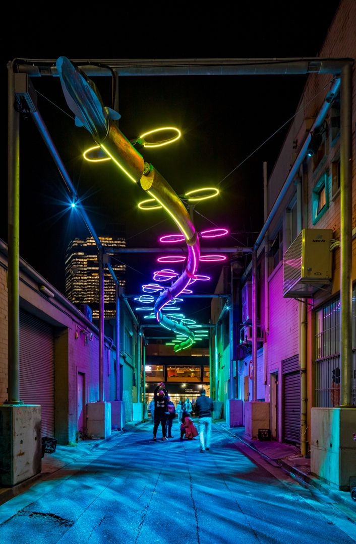 Neon-lit laneway at night with a glowing lizard-shaped light sculpture overhead and pedestrians walking beneath colorful illuminated rings.
