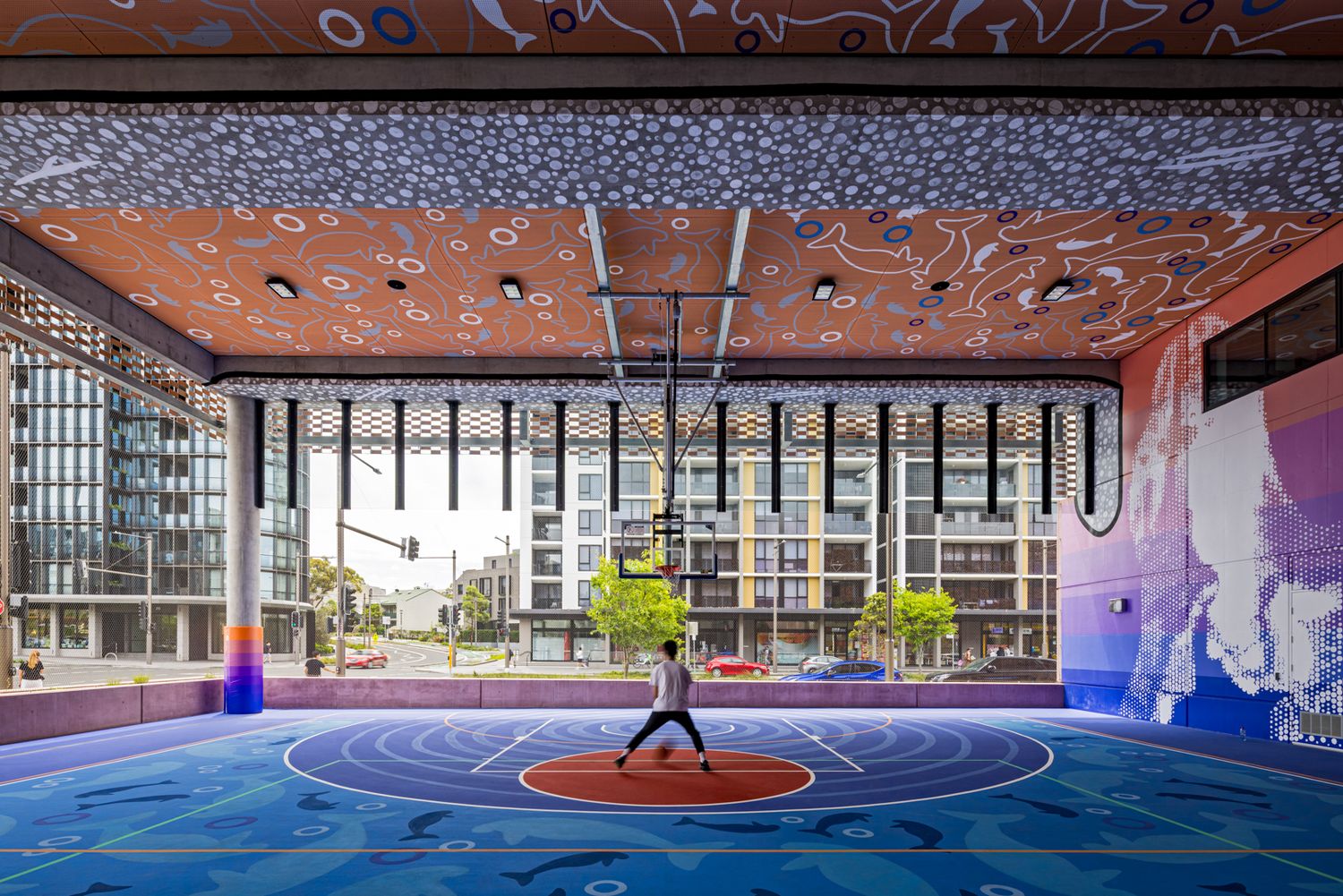 Covered urban basketball court with colorful painted flooring and murals as a person stands at center beneath a patterned ceiling.