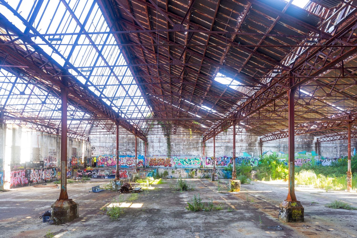 Abandoned industrial hall with a skylight roof, rusty steel columns, graffiti-covered walls and plants growing through the floor.