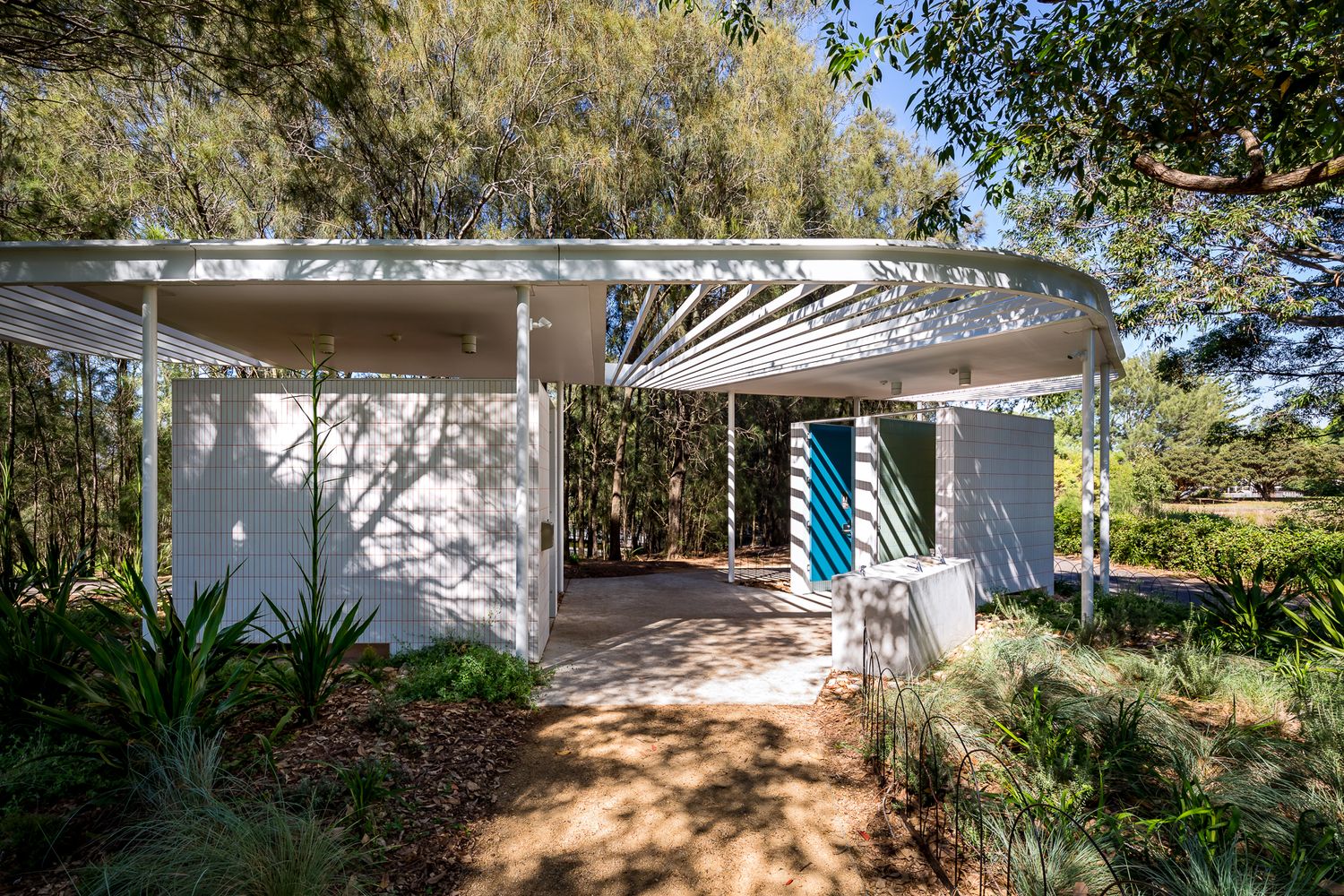 Aerial view of a curved white roof park shelter with teal doors, tiled walls and native plantings among eucalyptus trees.