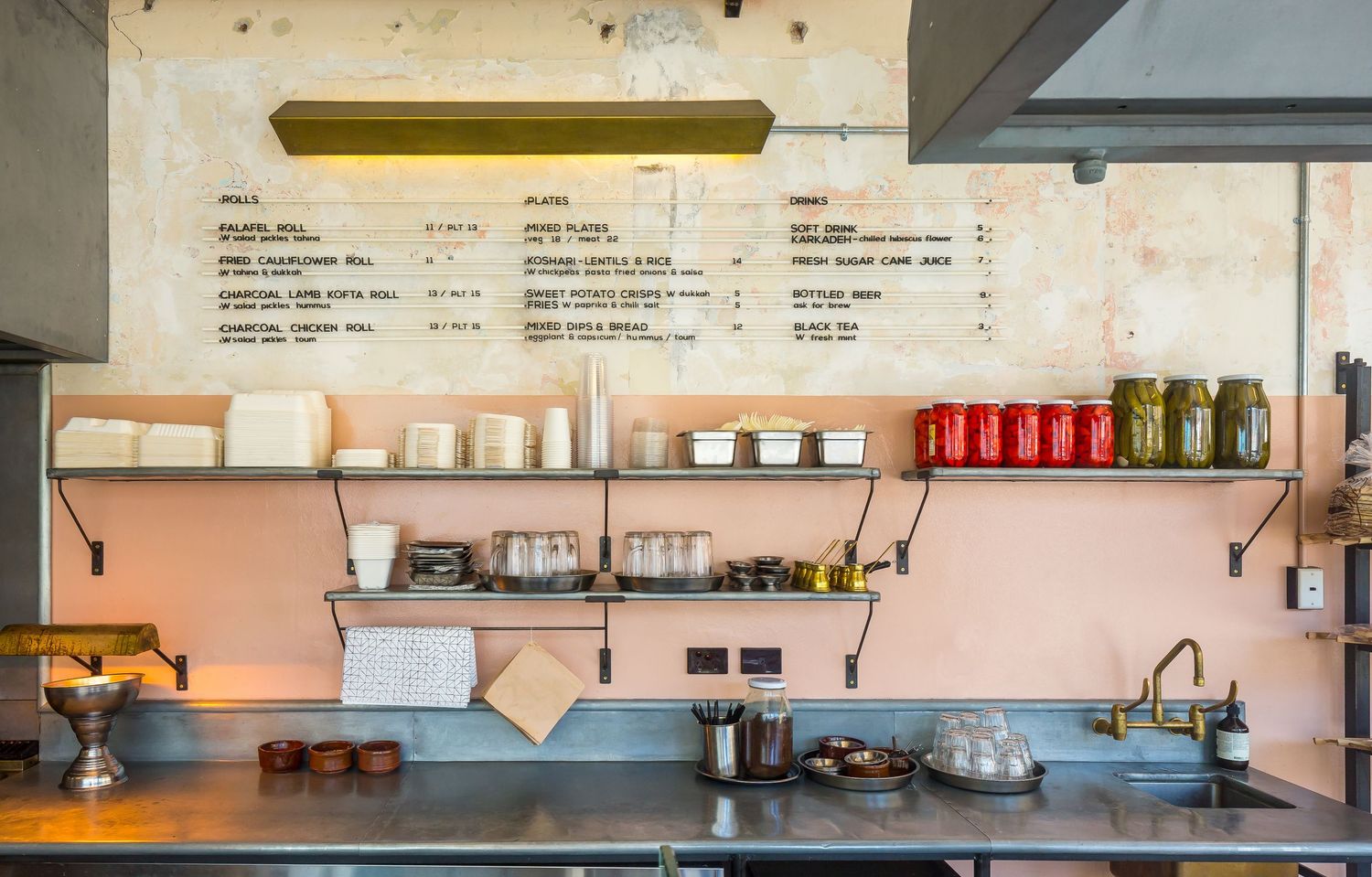 Cafe counter with a pink wall, wall-mounted menu board, open metal shelves holding jars, ceramics and cups above a stainless steel bench.