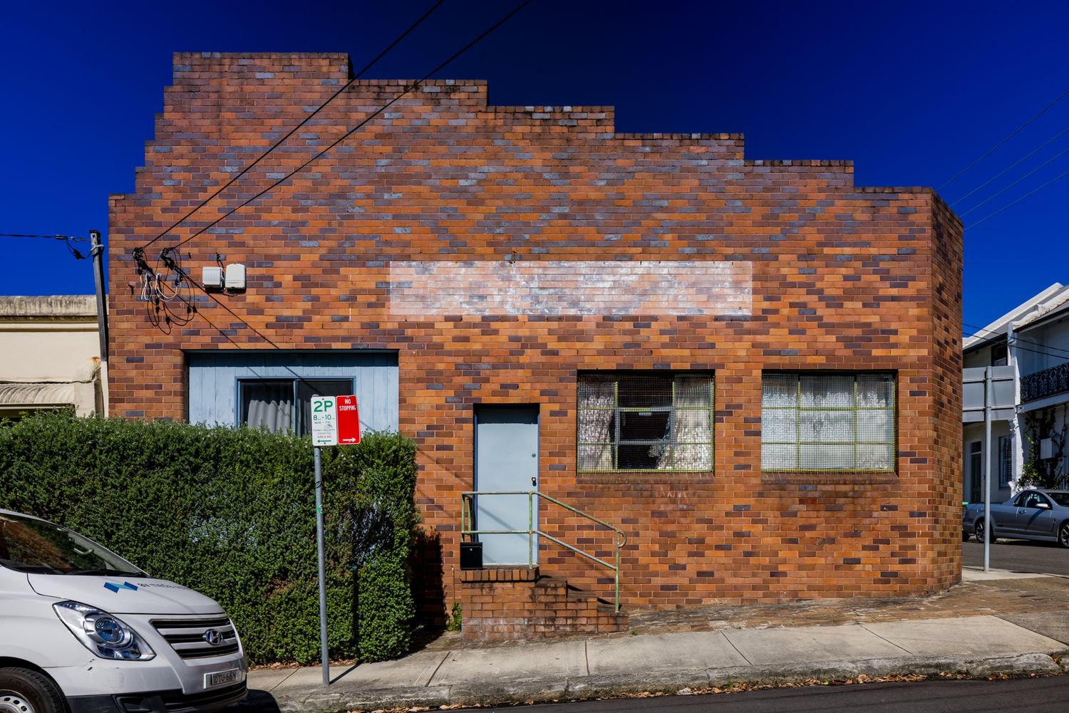 Small red brick commercial building with faded painted signage, boarded windows and a hedge on a quiet street corner.