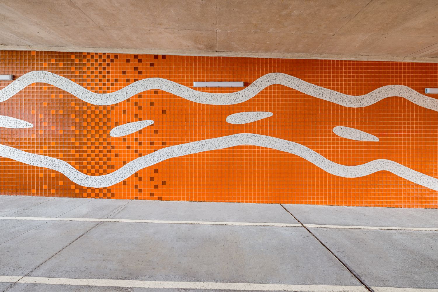 Bright orange tiled underpass wall with flowing white wavy lines and dotted patterns above a concrete pathway.