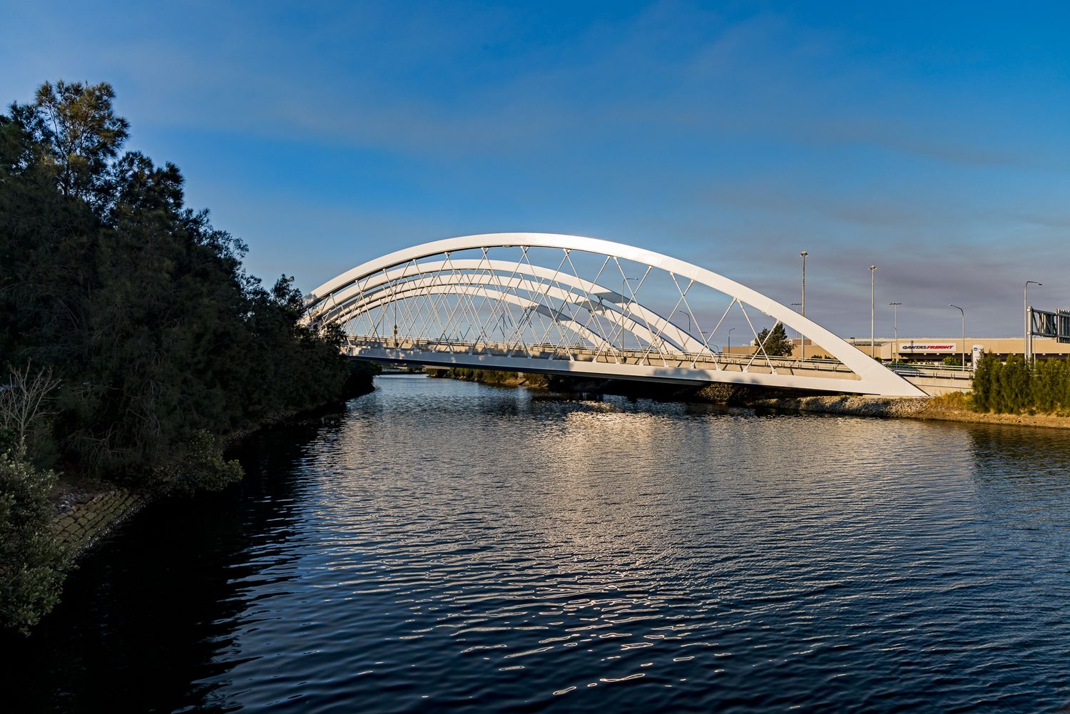 White steel arch bridge spanning a calm river with tree-lined banks under a blue evening sky.