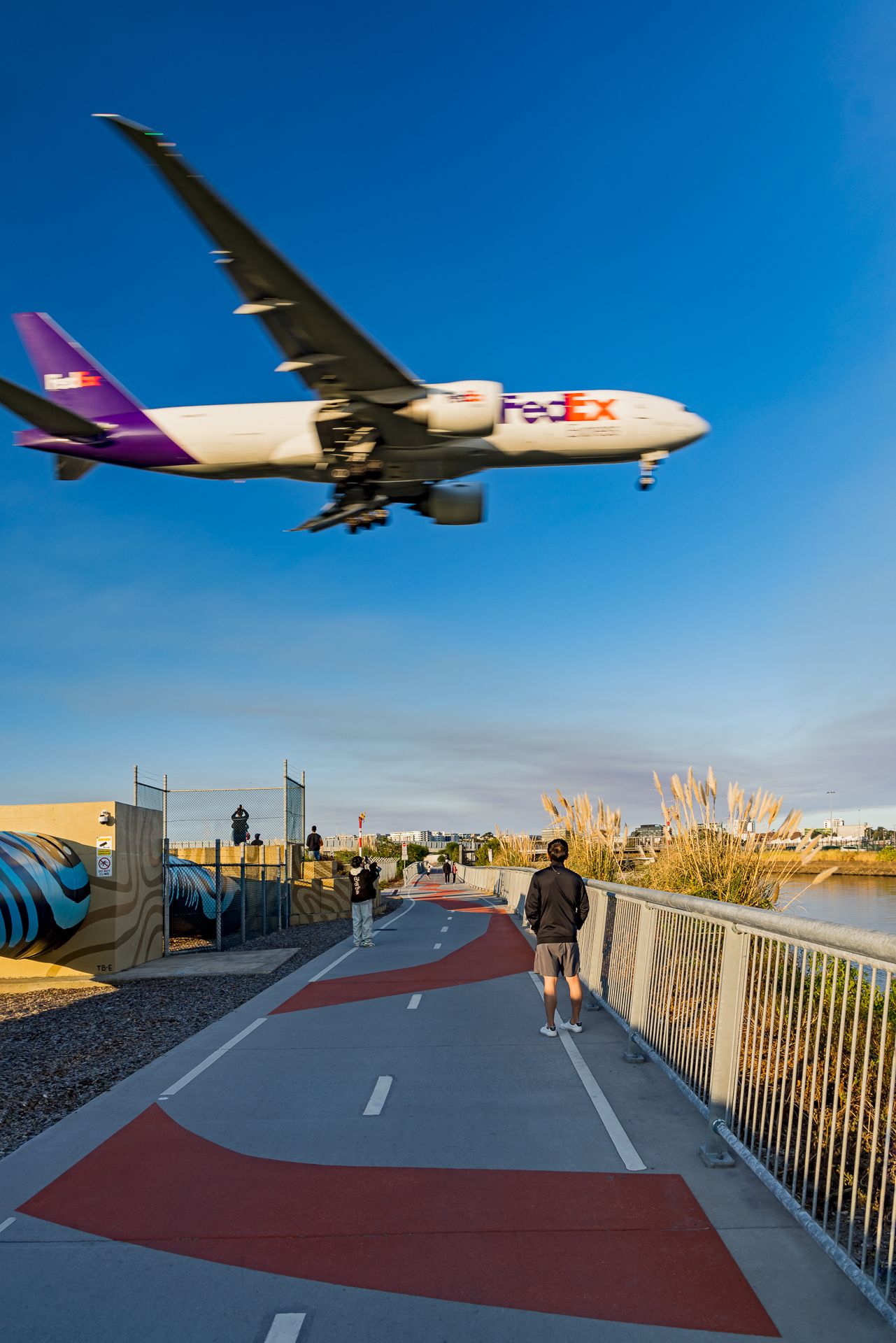 FedEx plane flying low over a red-marked cycle path as a man watches from beside a mural wall and railing.