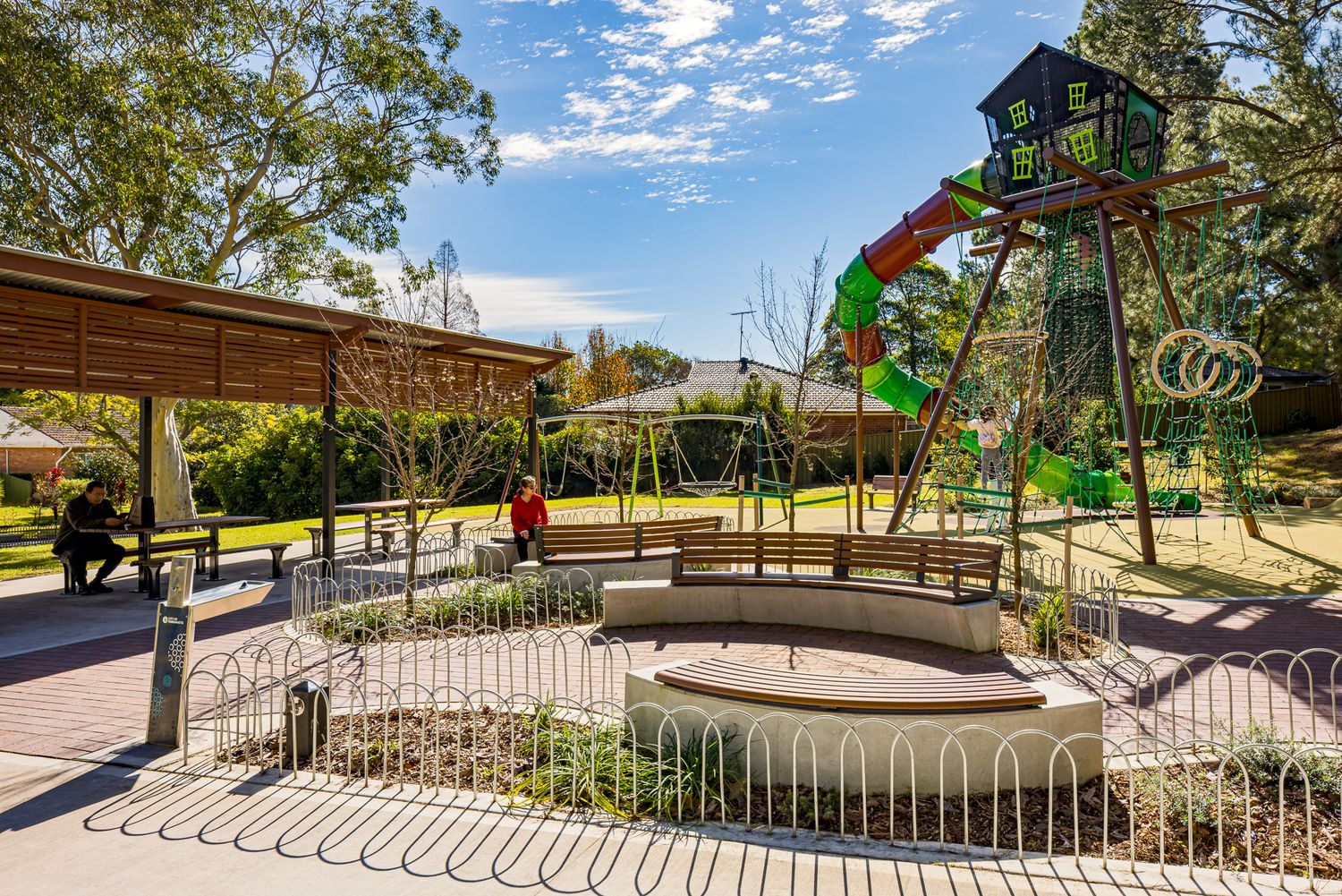 Children's playground with a treehouse-style climbing tower and slide beside a shaded pavilion and curved concrete seating in a leafy park.