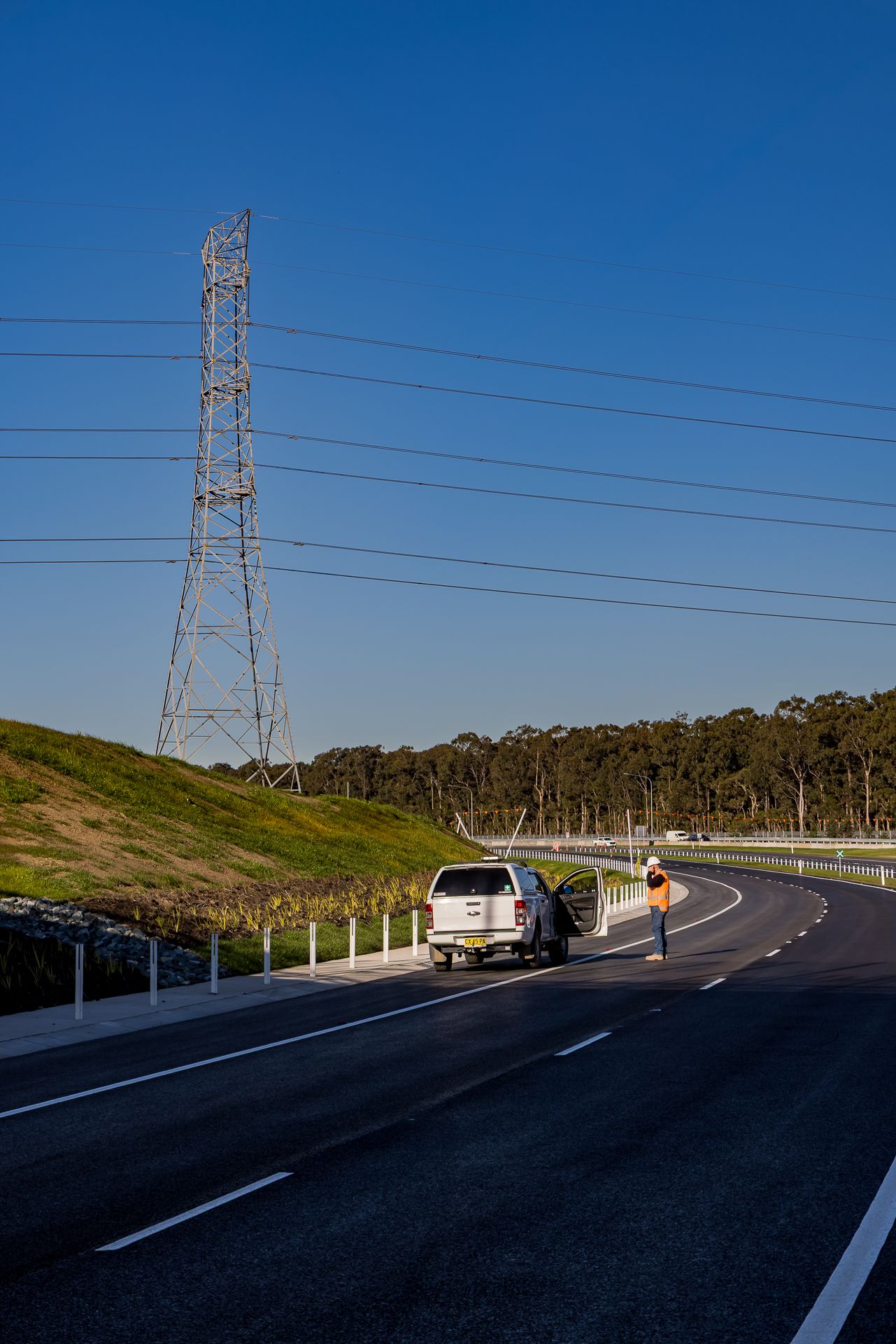 Worker in high-visibility gear stands beside a white utility vehicle on a new road near a tall electricity pylon.