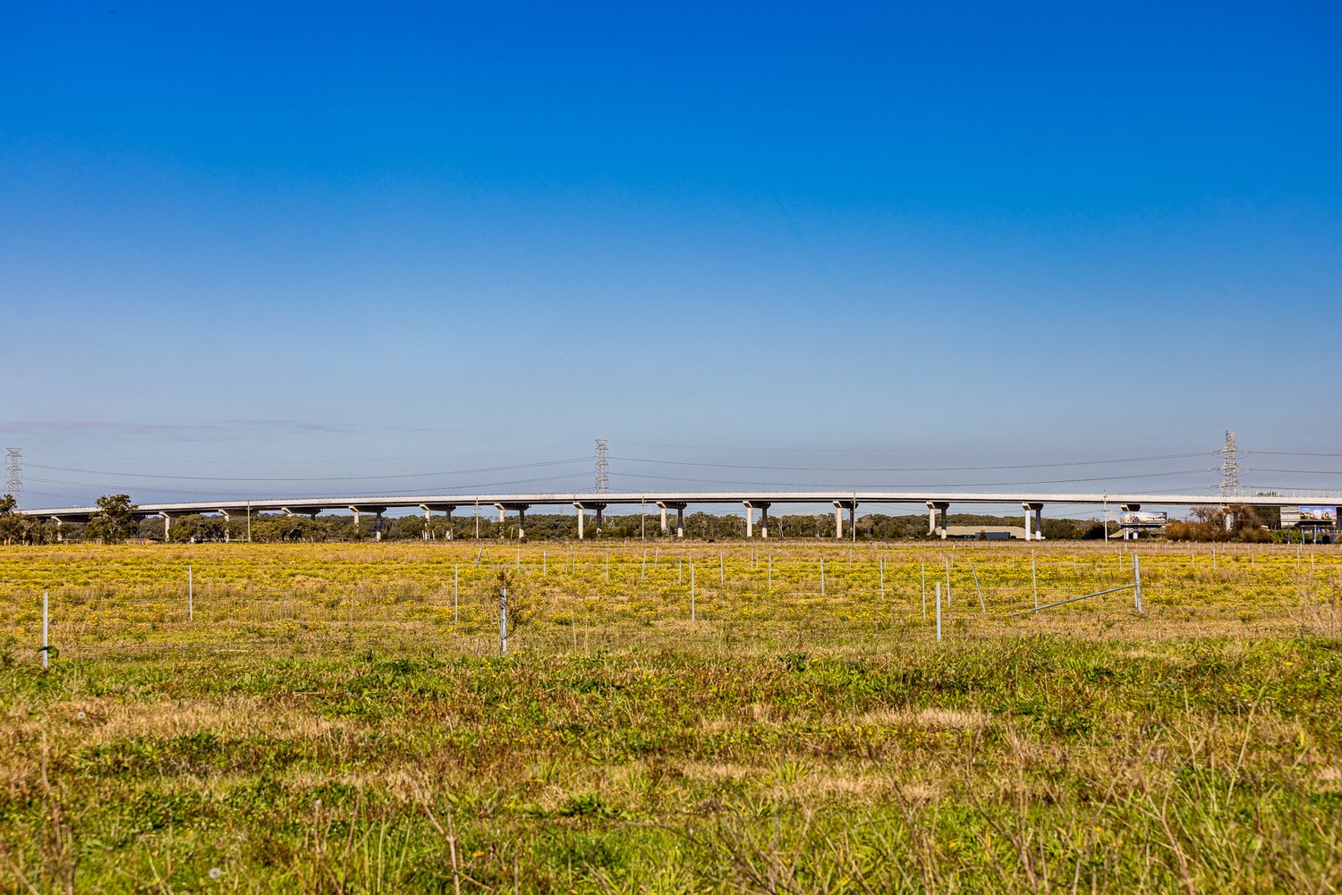 Long elevated road bridge crossing a flat yellow-green field under a wide, clear blue sky.