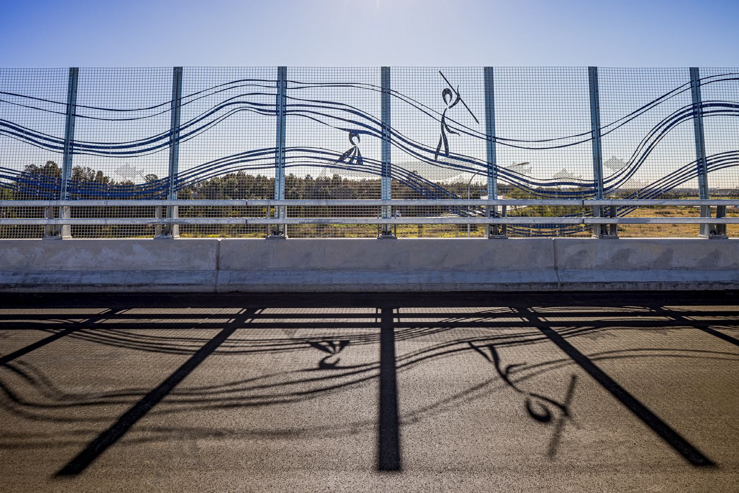 Artistic bridge barrier with wave patterns and dancing figure designs casting long shadows across the sunlit road deck.