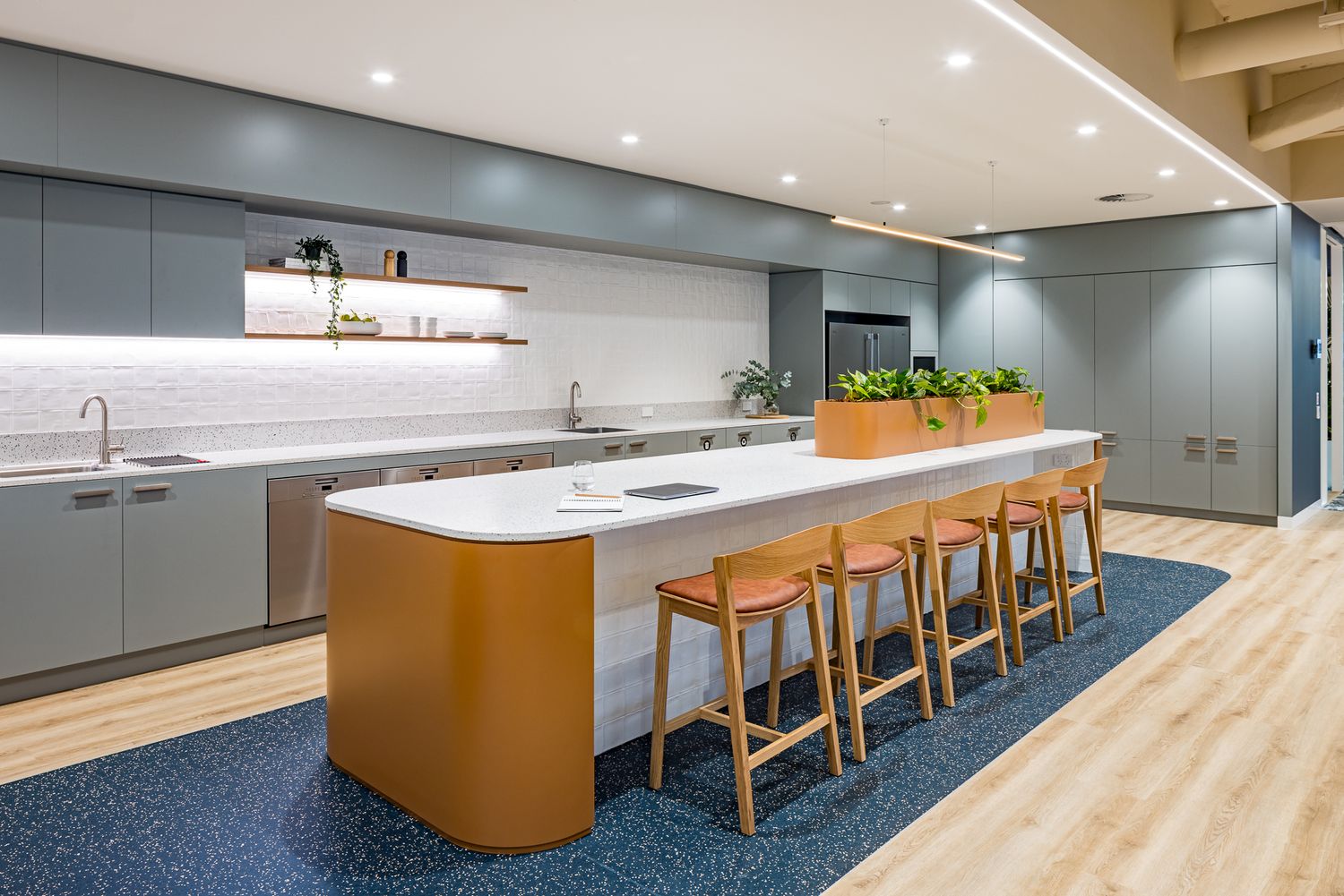 Sleek office kitchen with a curved white island, wooden bar stools, gray cabinets, a built-in planter and blue patterned carpet.