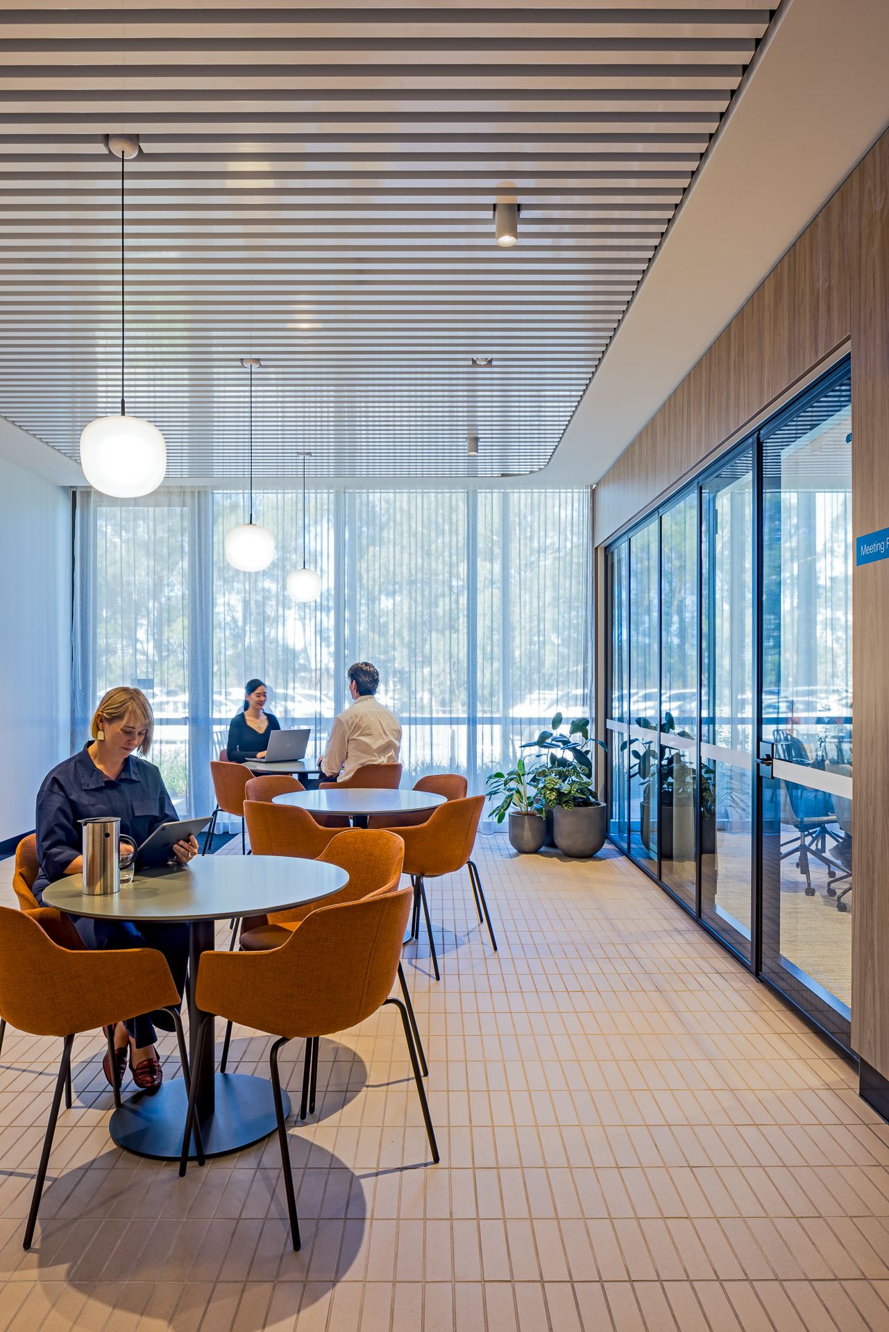Bright cafe-style break area with orange chairs, round tables and pendant lights beside floor-to-ceiling windows and glass-walled offices.