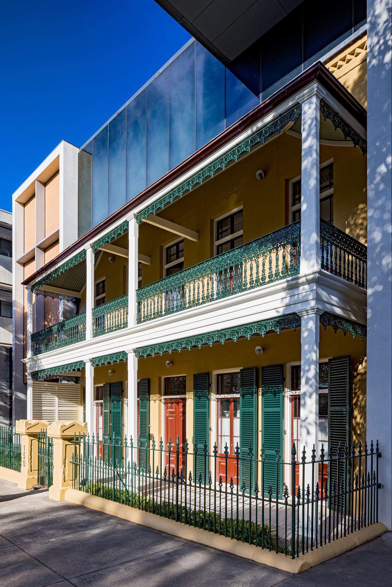 Historic two-storey terrace with yellow walls, green cast-iron lace balconies and white columns beside a modern glass building.