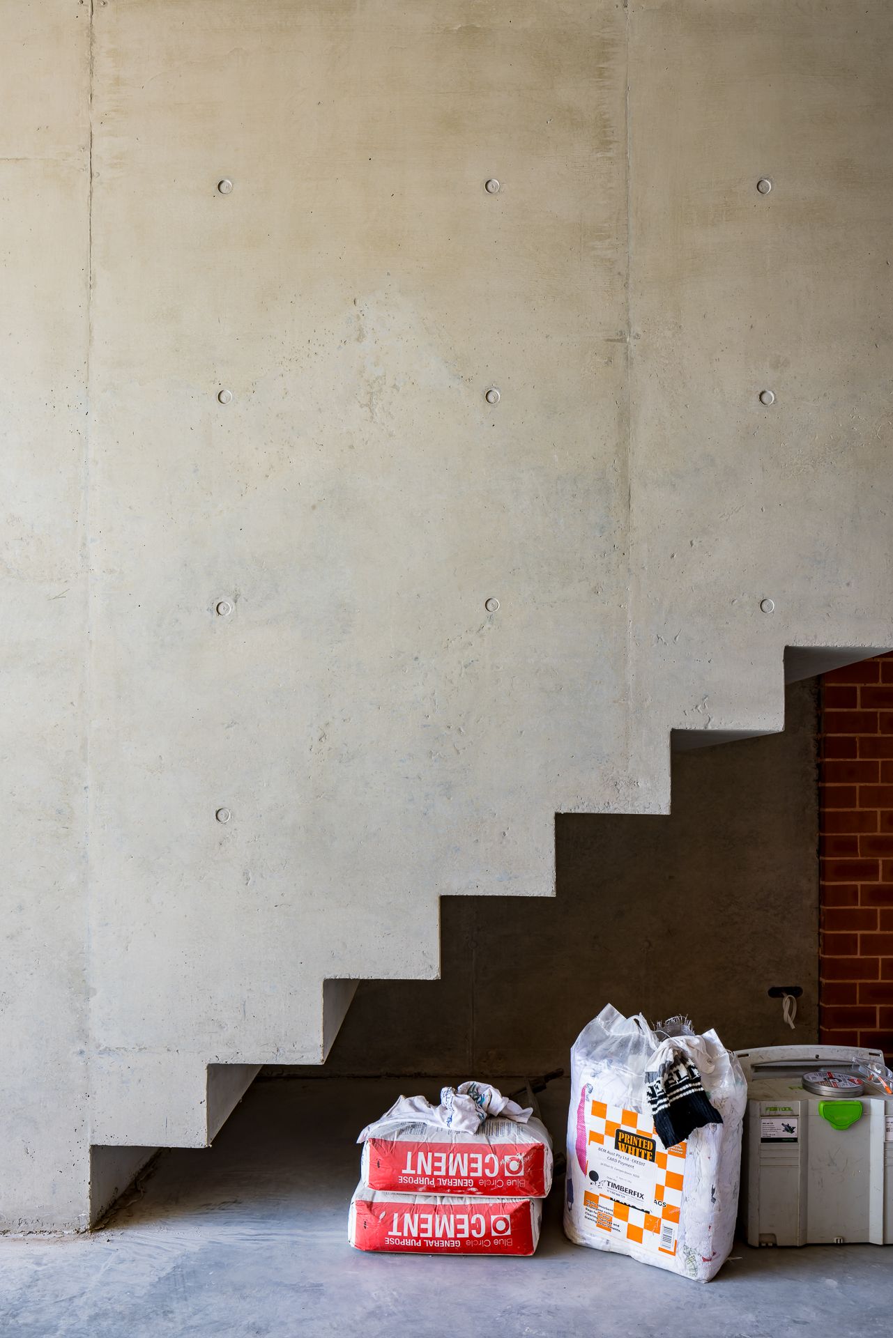 Exposed concrete wall with a floating staircase above bags of cement and a dust extractor on the floor of a building site.