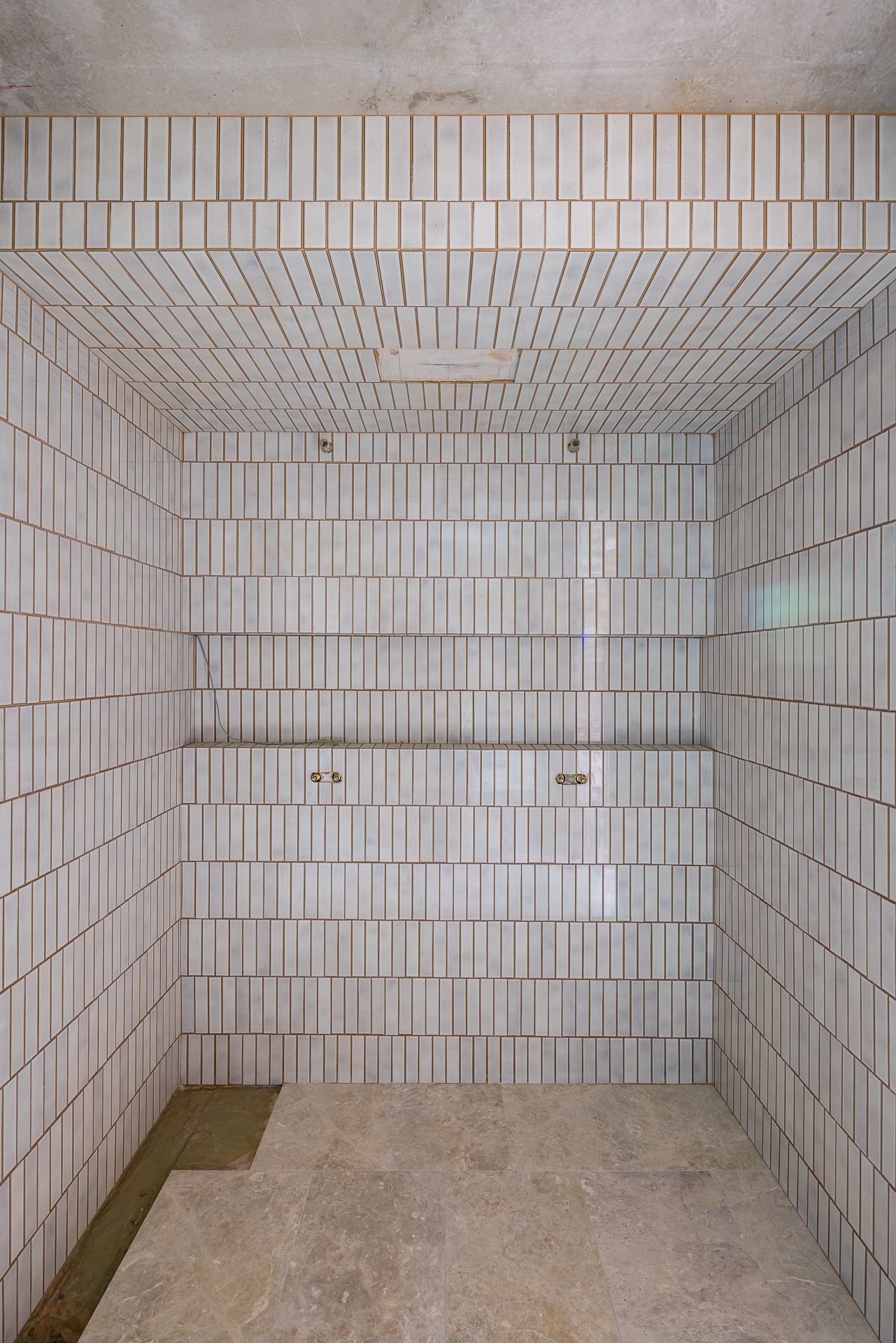 Empty shower recess lined with narrow vertical white tiles on the walls and ceiling, with a stone-look floor and two wall fixtures.