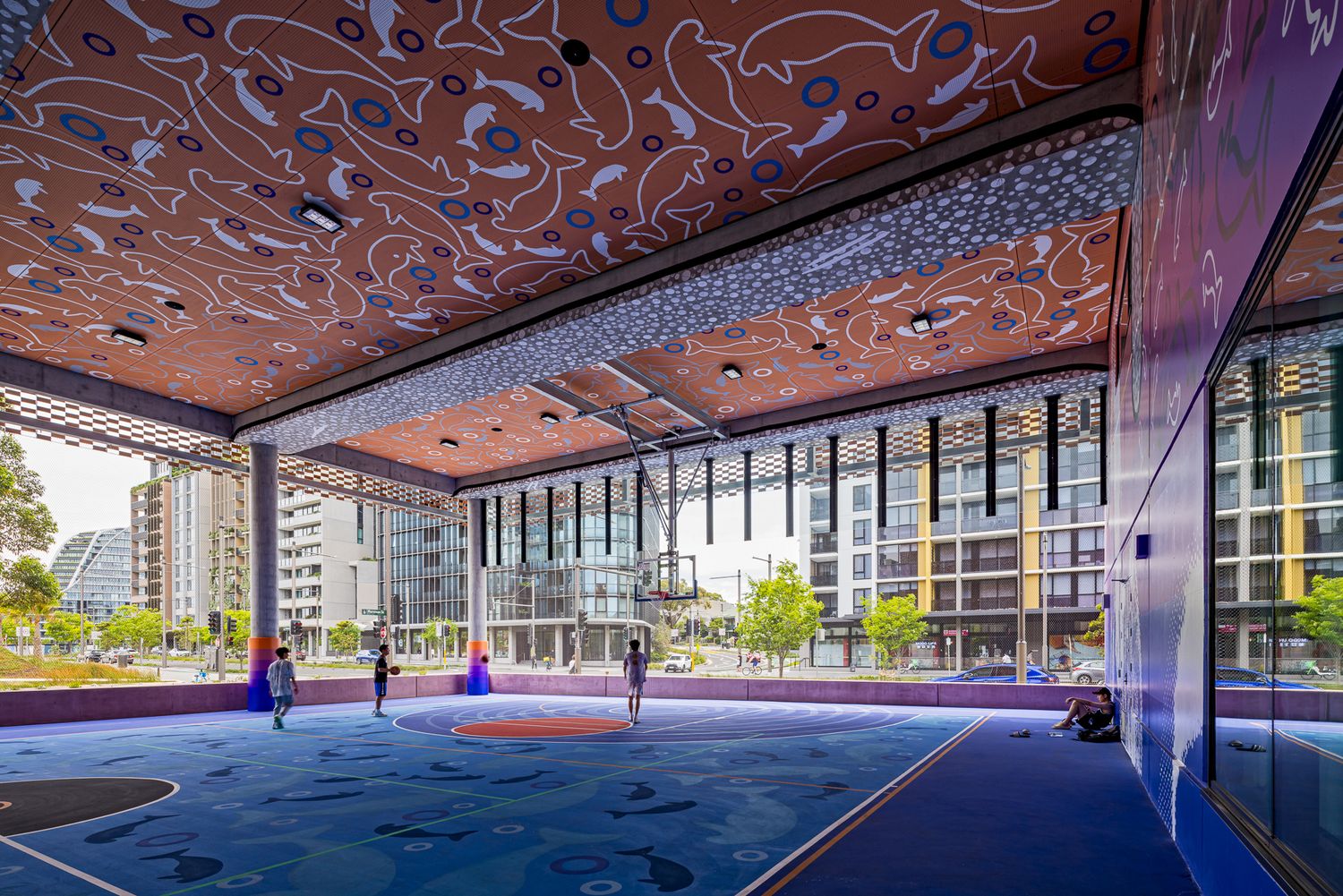 Rooftop basketball court with a colorful blue floor and an ornate patterned ceiling overlooking high-rise buildings.
