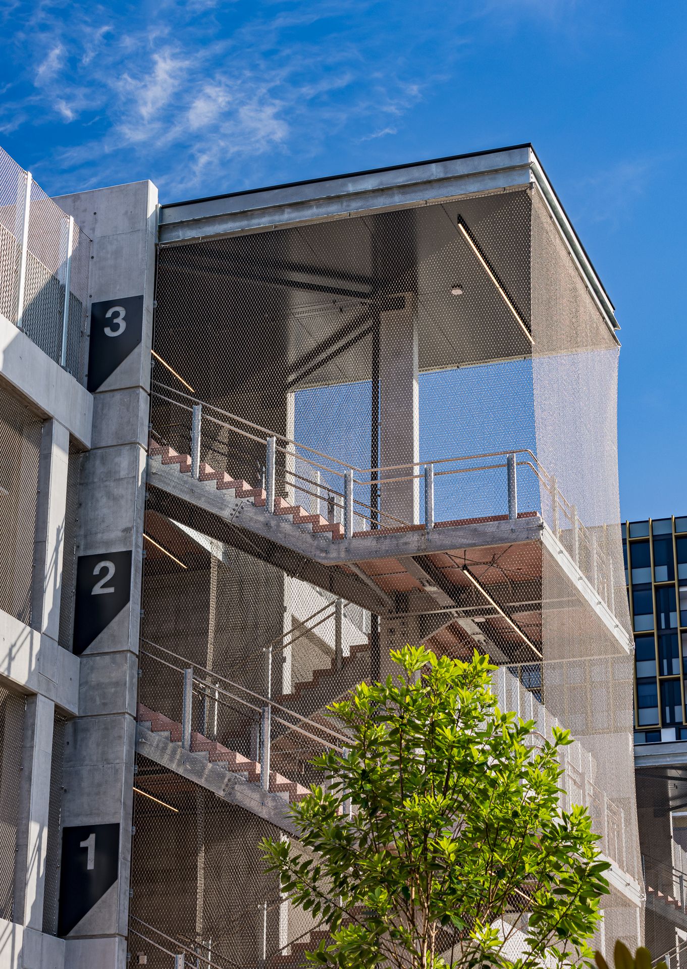 Multi-level concrete parking structure stairs with numbered levels one to three, mesh screening and a green tree in front.