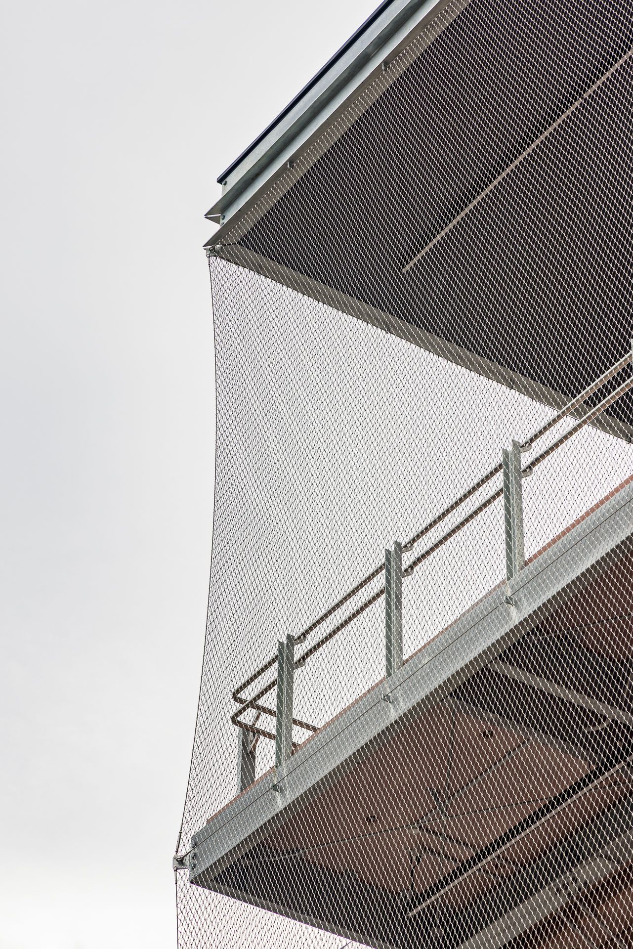 Upward view of a modern multi-level building with a stainless steel mesh facade and metal railings against a pale gray sky.