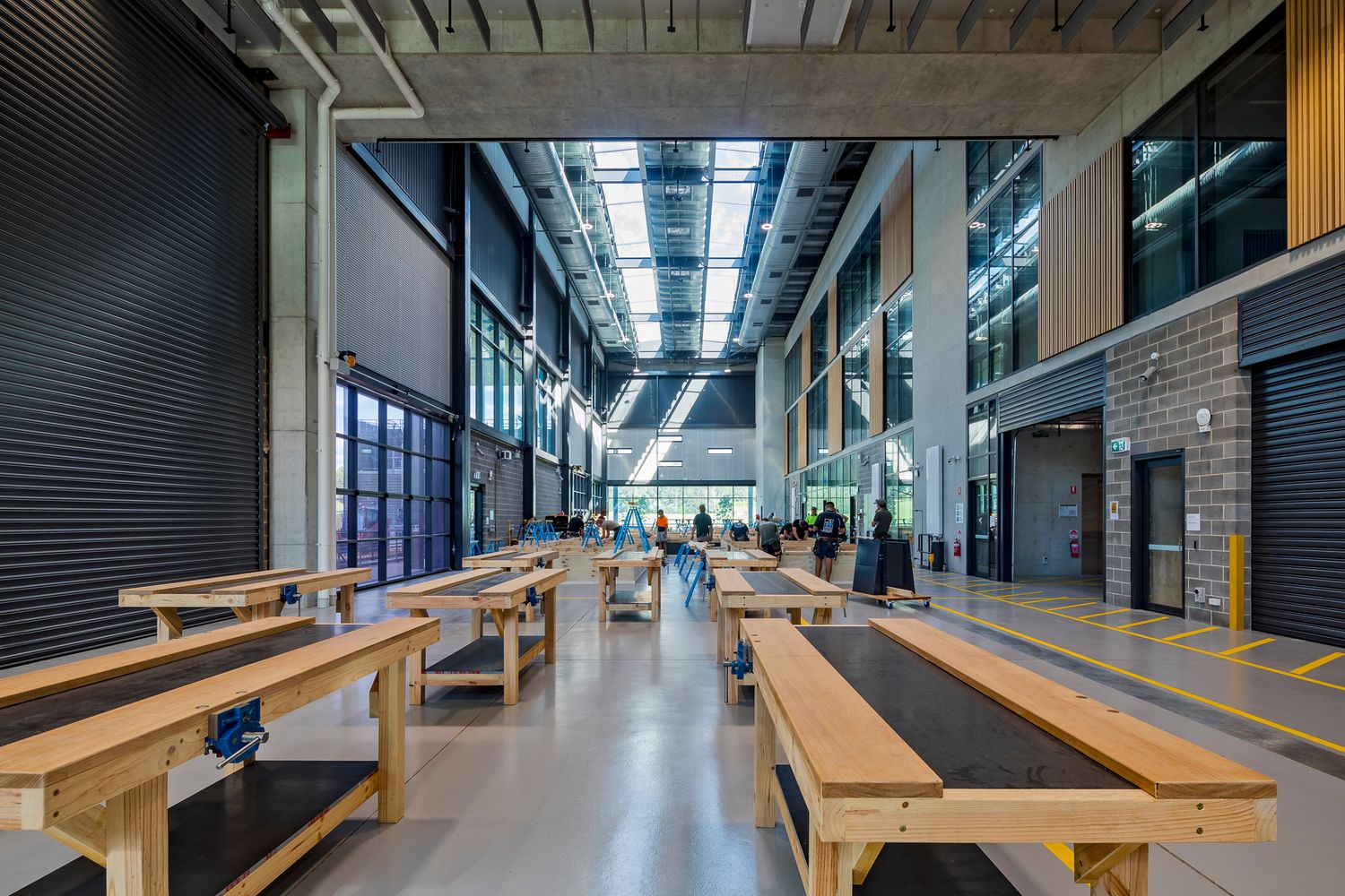 Spacious TAFE workshop hall with timber workbenches, roller doors, high skylights and students working in the background.