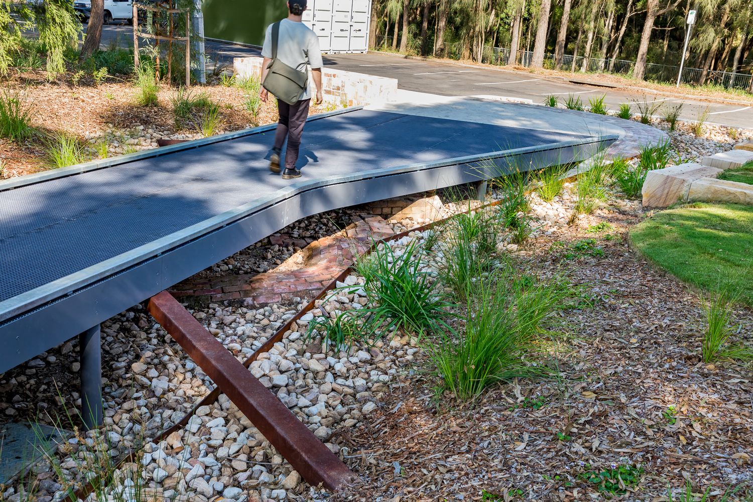 A pedestrian walks along an elevated metal boardwalk over pebble beds, grasses and a rusted steel beam in a park.